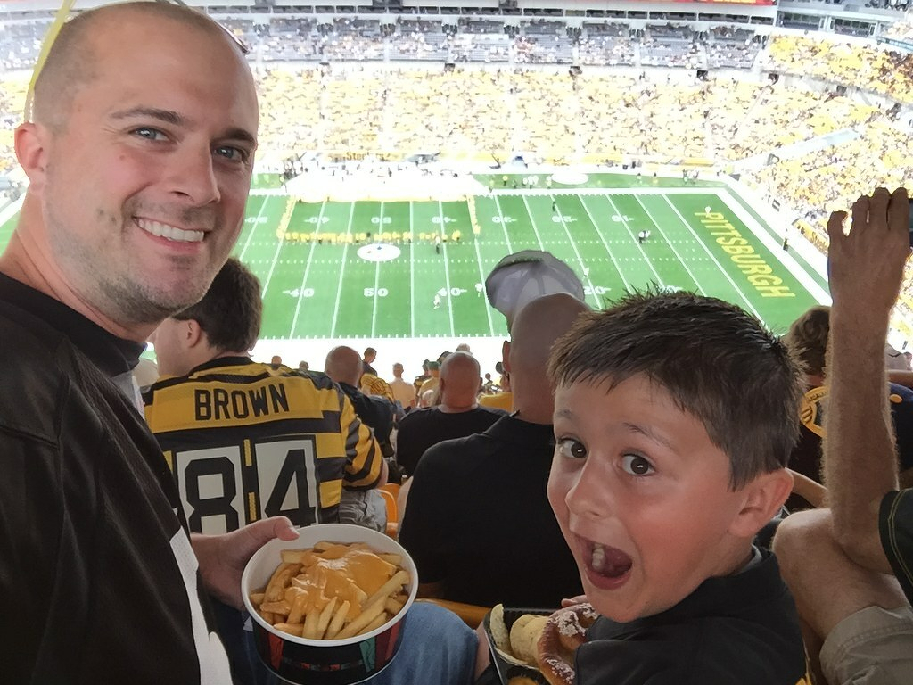 A man and a child are at a football game, eating snacks and surrounded by people wearing team jerseys.