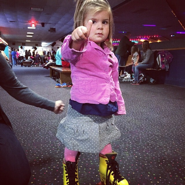 A child wearing a pink jacket and roller skates is giving a thumbs-up in a roller skating rink with other people in the background.