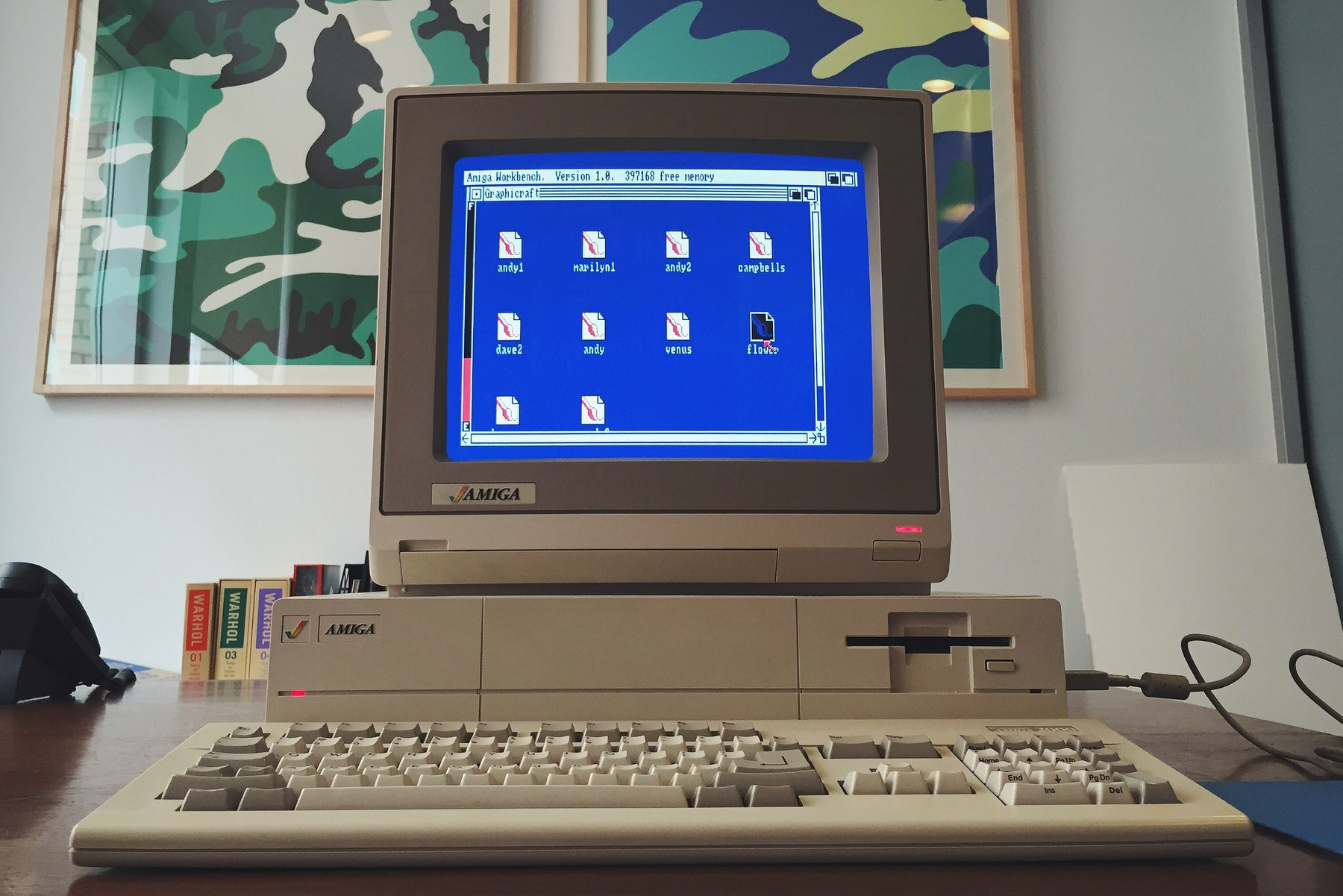 A vintage Amiga computer setup is displayed on a desk with colorful camouflage artwork on the wall behind it.