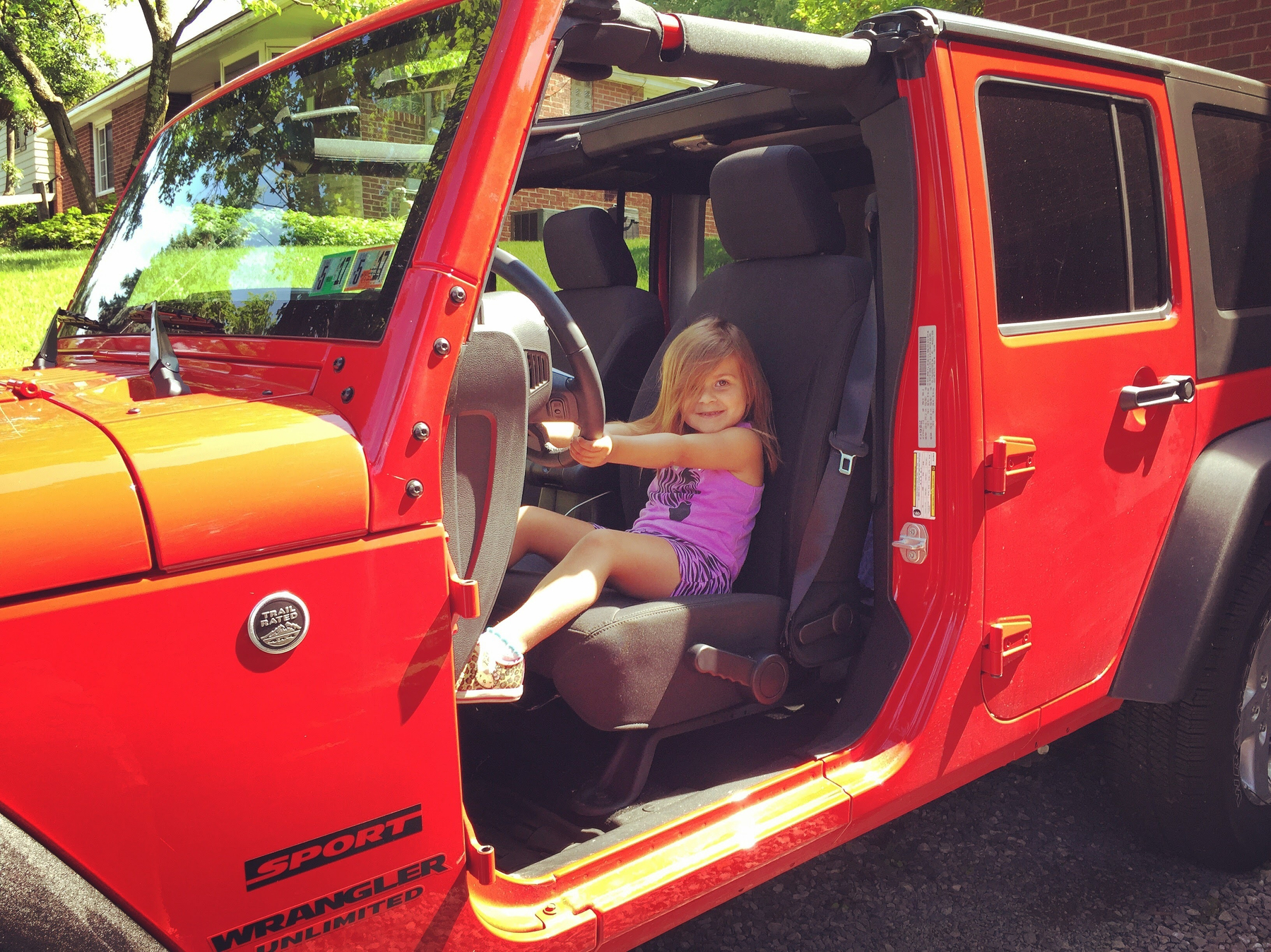 A young girl is sitting in the driver's seat of a bright orange Jeep Wrangler with the door open.