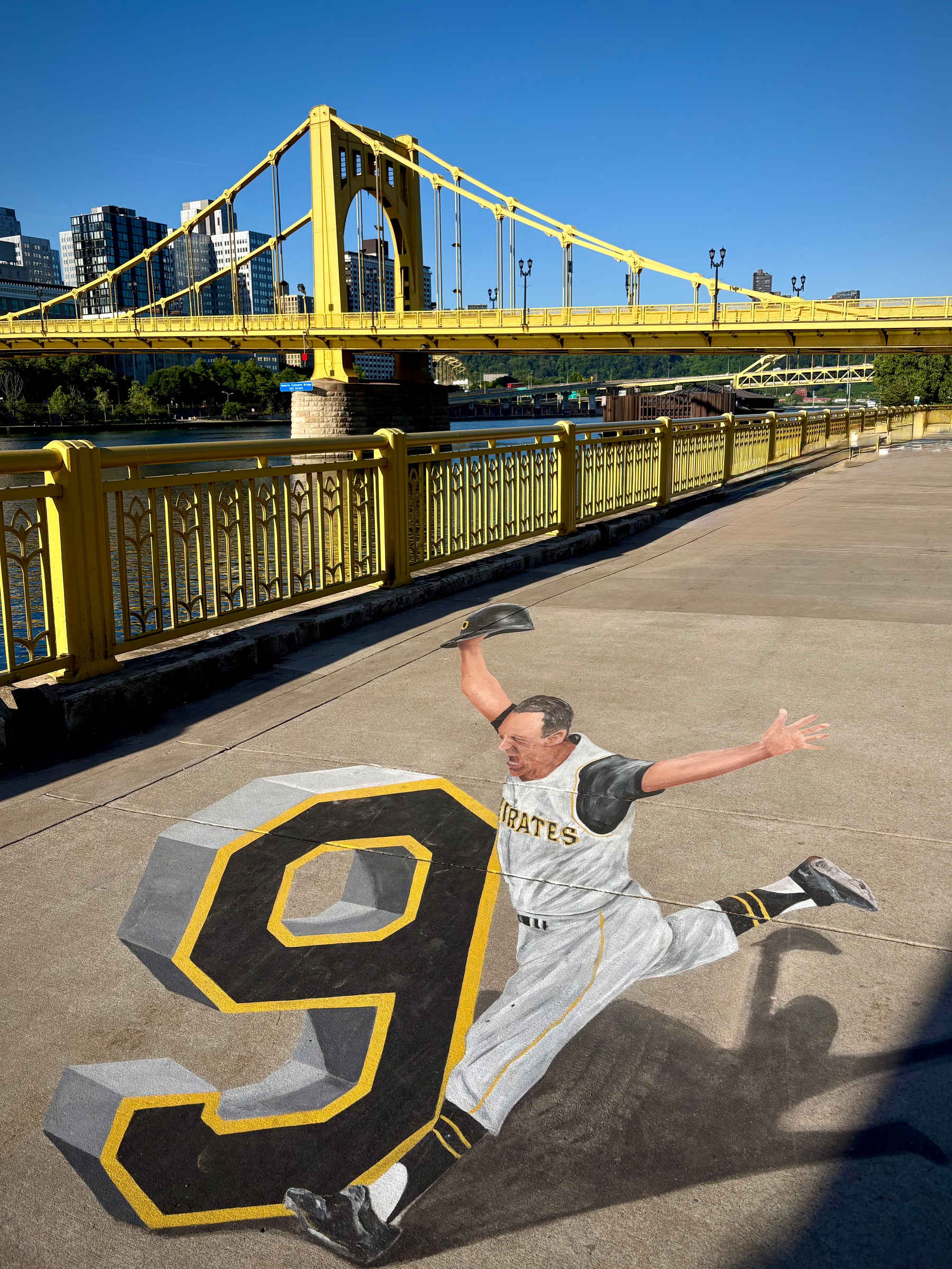 A 3D street painting depicts a baseball player in mid-slide beside a large number nine on a riverside walkway with a yellow bridge and city buildings in the background.