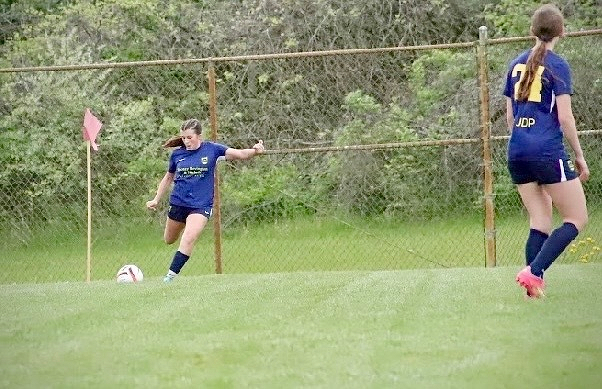 Two soccer players in blue uniforms are on a grass field, with one kicking a ball near a corner flag.