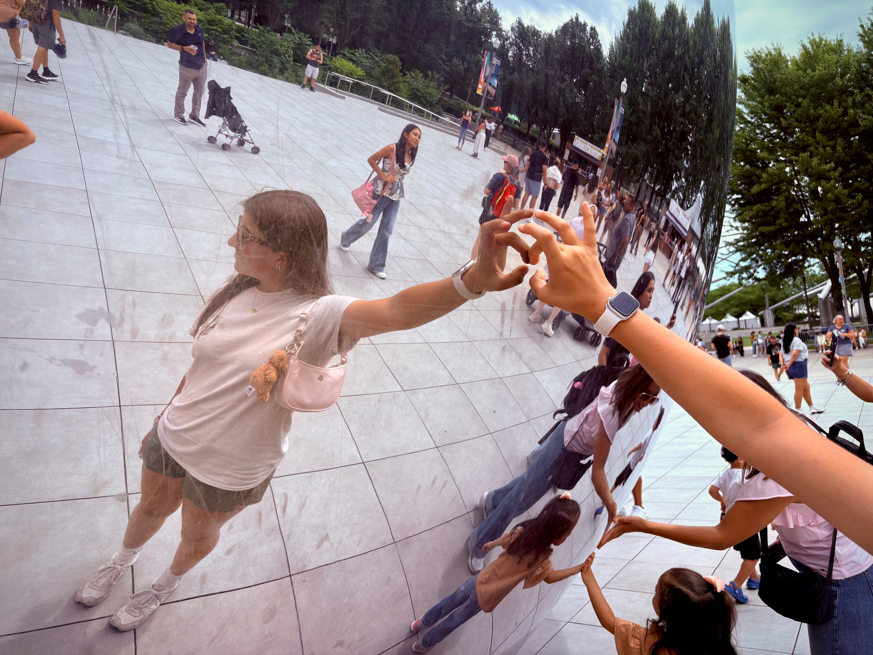 A person touches the reflective surface of a large, curved metallic sculpture, creating mirrored reflections, while people walk around in an outdoor setting.