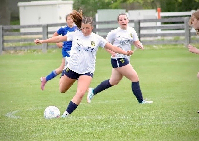 A soccer player in a white jersey is about to kick the ball during a match, with teammates and opponents visible in the background on a grass field.