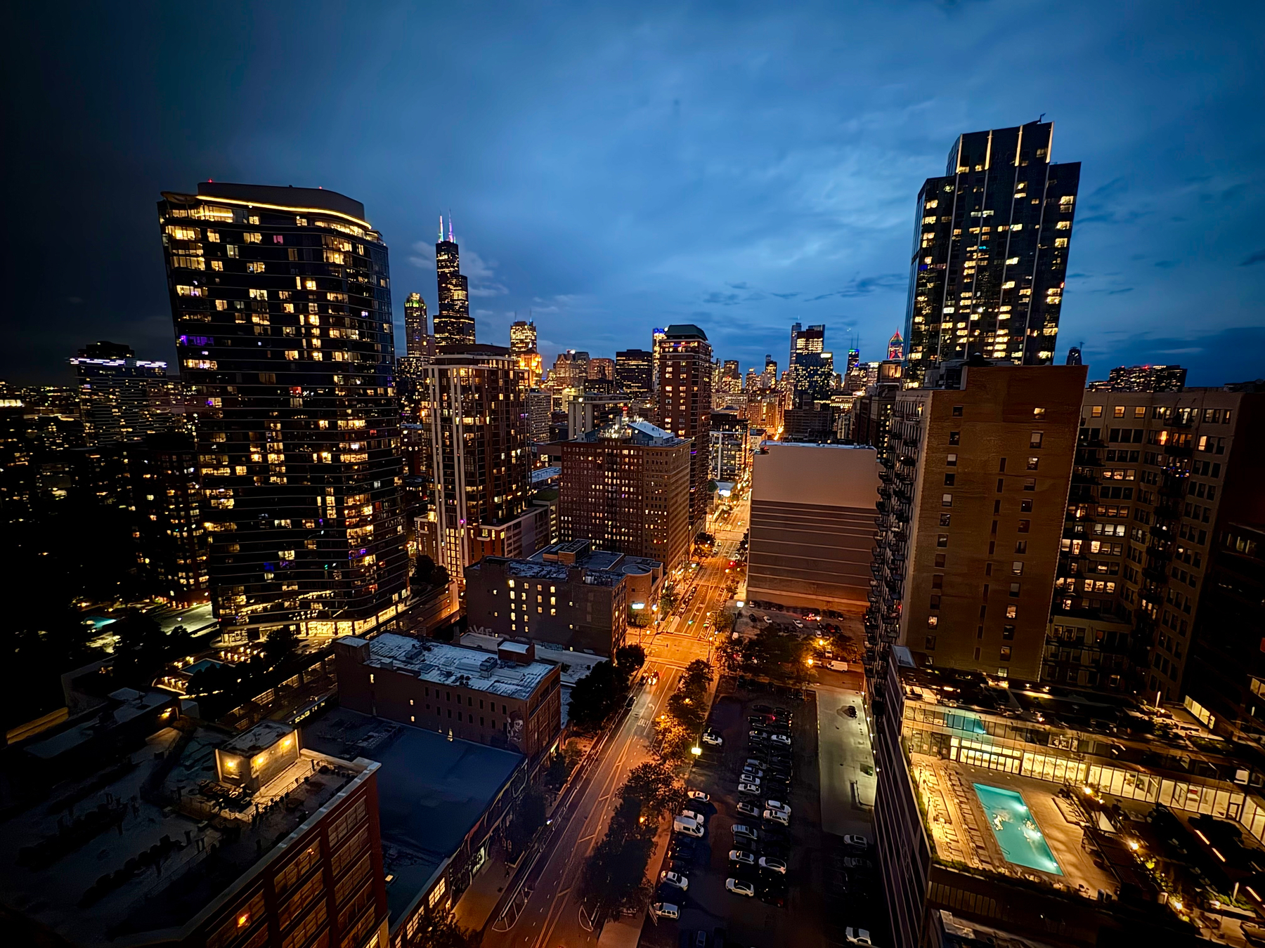 A bustling cityscape at twilight features illuminated skyscrapers and a vibrant street scene with glowing lights and a rooftop pool.