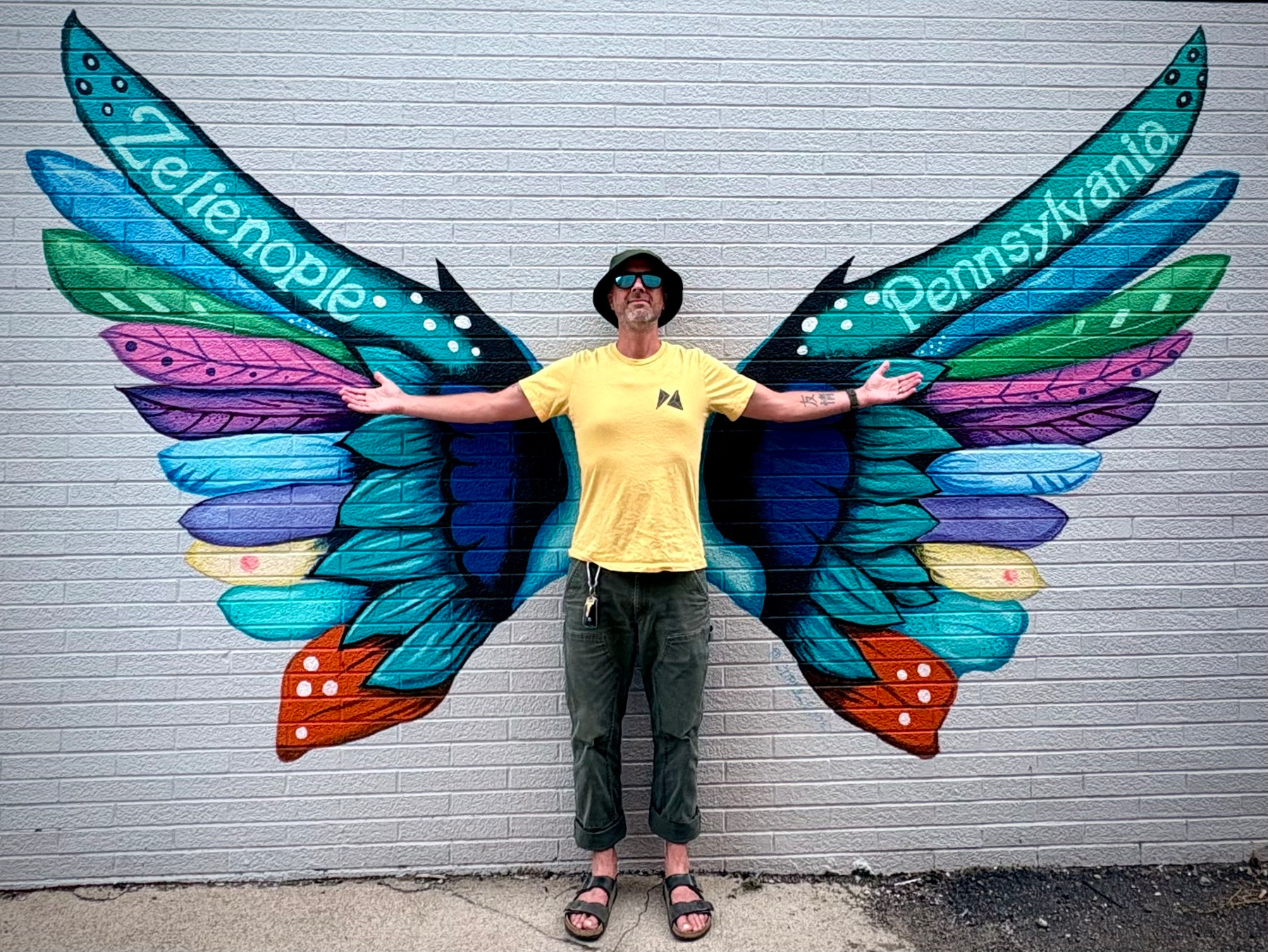 A person stands with arms outstretched in front of a colorful butterfly mural painted on a wall, with the words Zelienople and Pennsylvania on the wings.