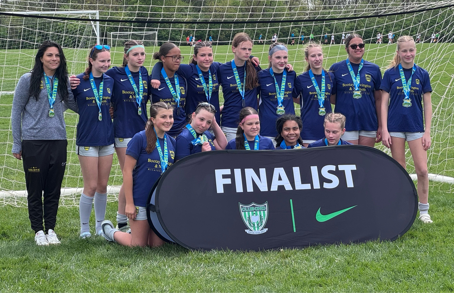 A youth soccer team poses with medals and a Finalist banner in front of a goal on a grassy field.