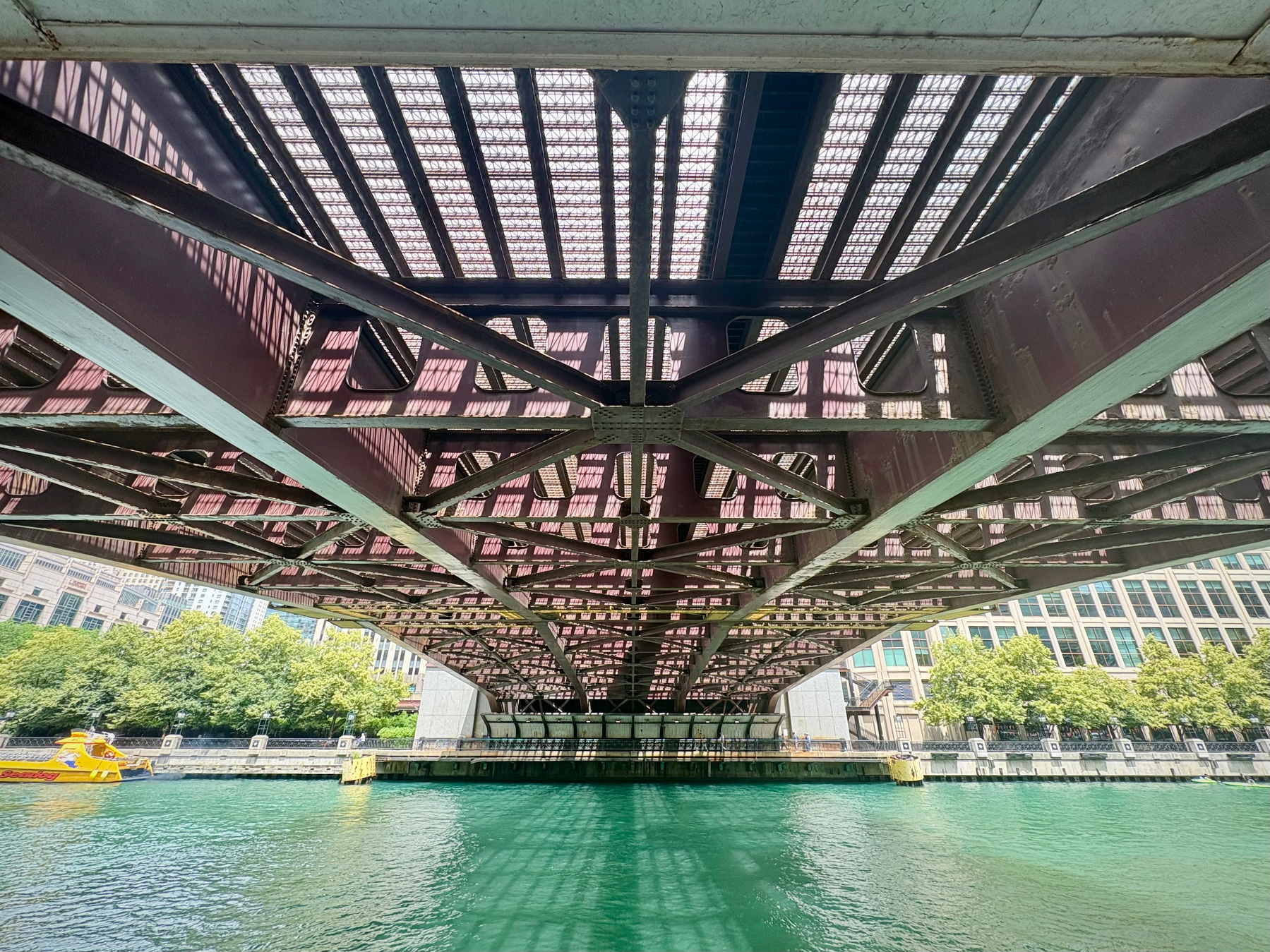 A view from beneath a bridge shows its metal framework spanning a clear, greenish-blue body of water, flanked by urban buildings and trees on the shore.