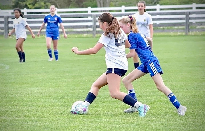 Two soccer players in a match are competing for control of the ball while other players observe in the background on a grass field.