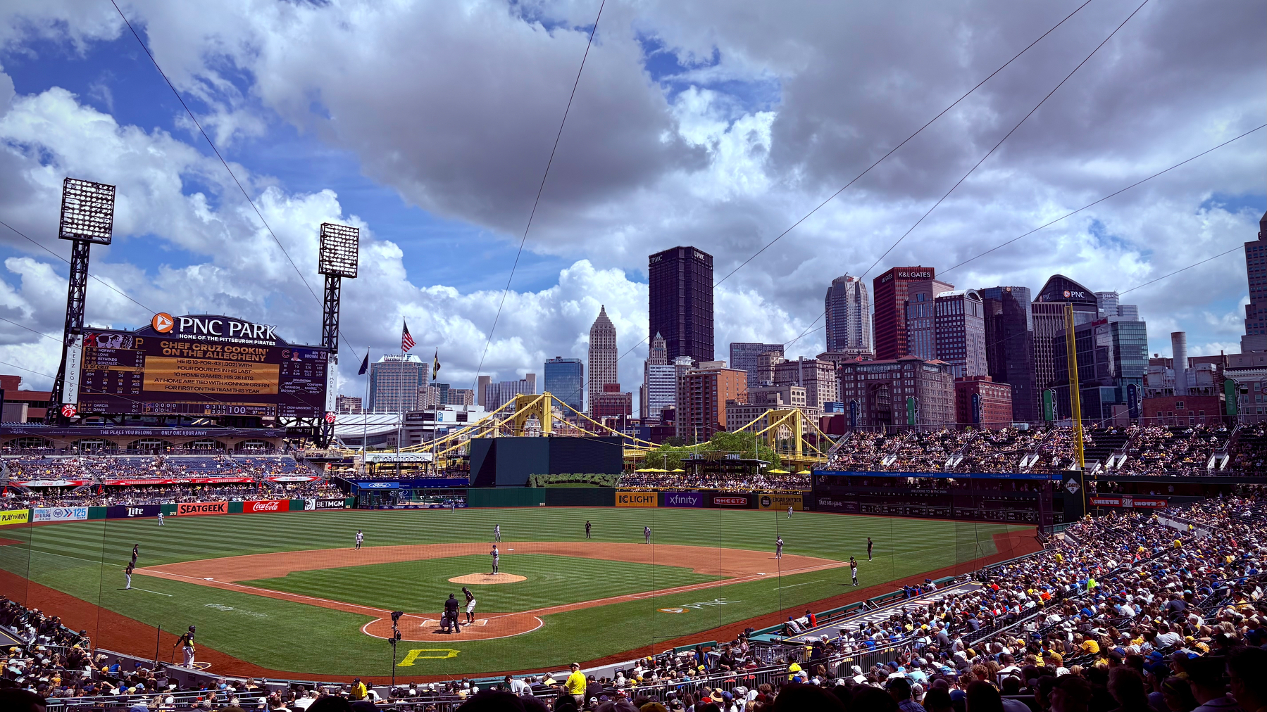 A baseball game is taking place at a stadium with a city skyline and bridges in the background under a partly cloudy sky.