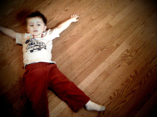 A young child wearing a white shirt and red pants is lying on a wooden floor with arms outstretched.