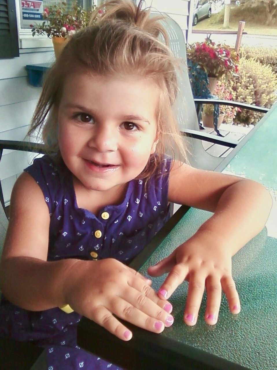 A young girl with painted nails and a purple dress smiles while seated at a glass table on a porch.