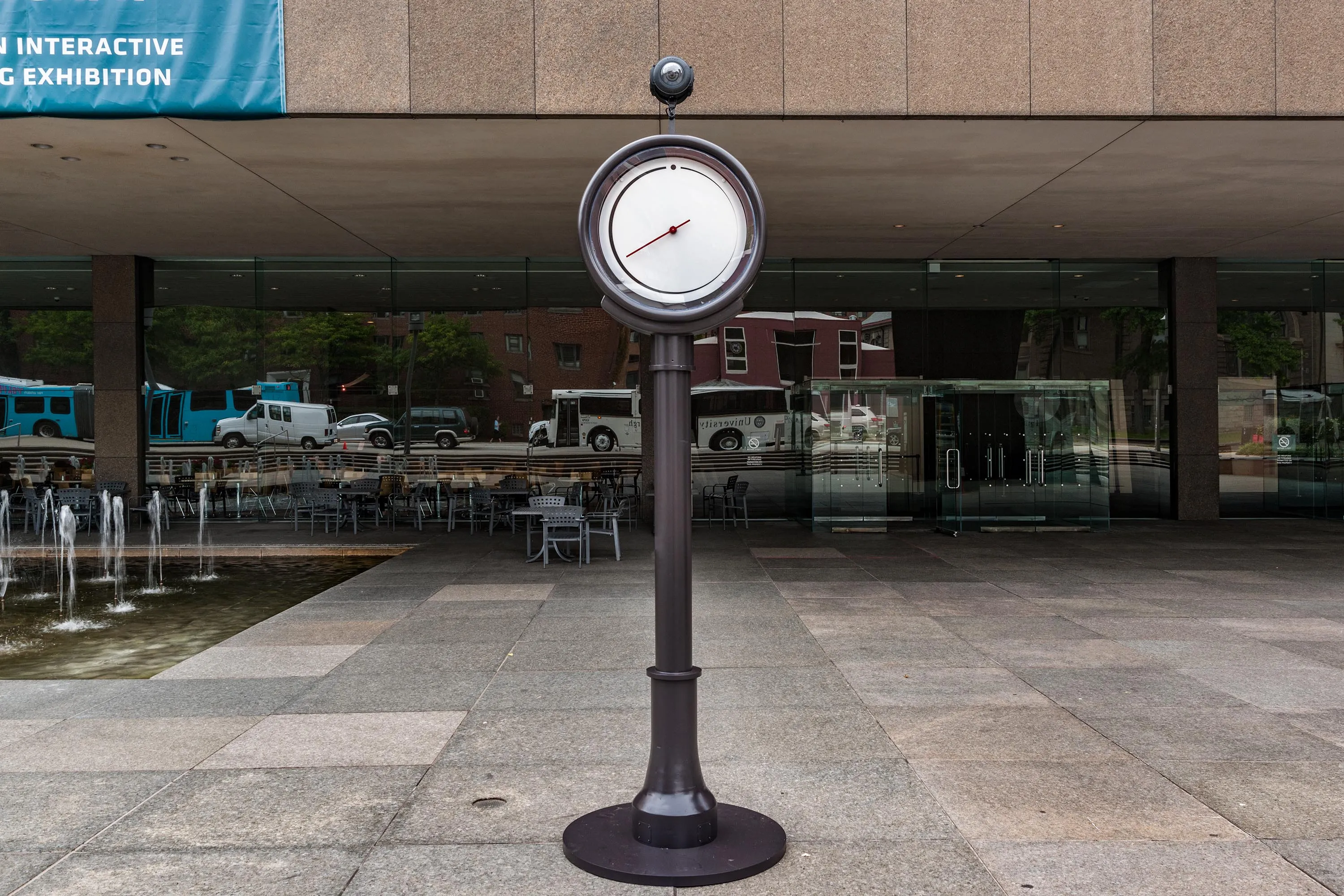 Auto-generated description: A large outdoor scale with a round dial is positioned on a plaza in front of a building with large glass windows.
