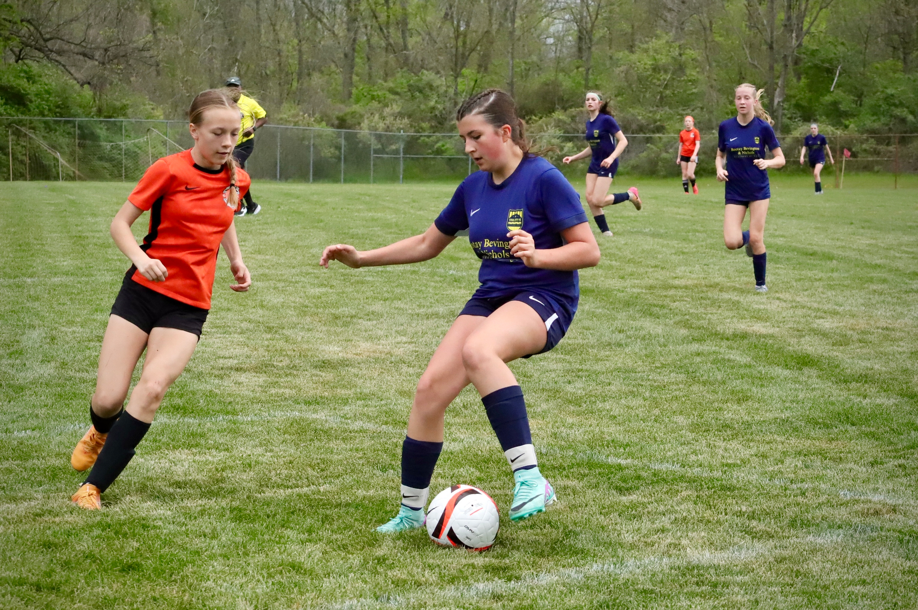 Two young soccer players are competing for the ball on a grassy field, while other players and a referee are visible in the background.