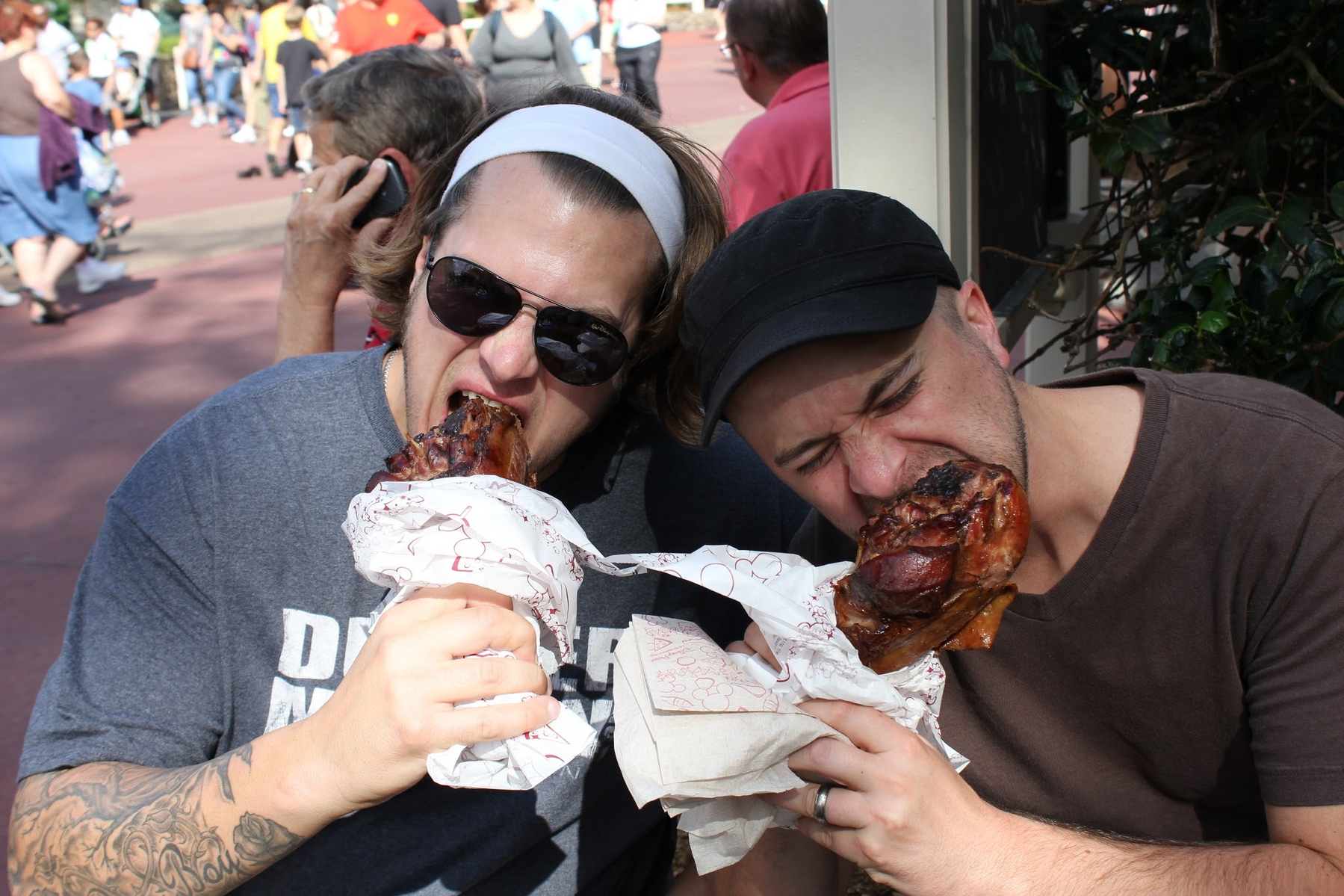 Two people are enthusiastically eating large turkey legs while sitting outdoors.