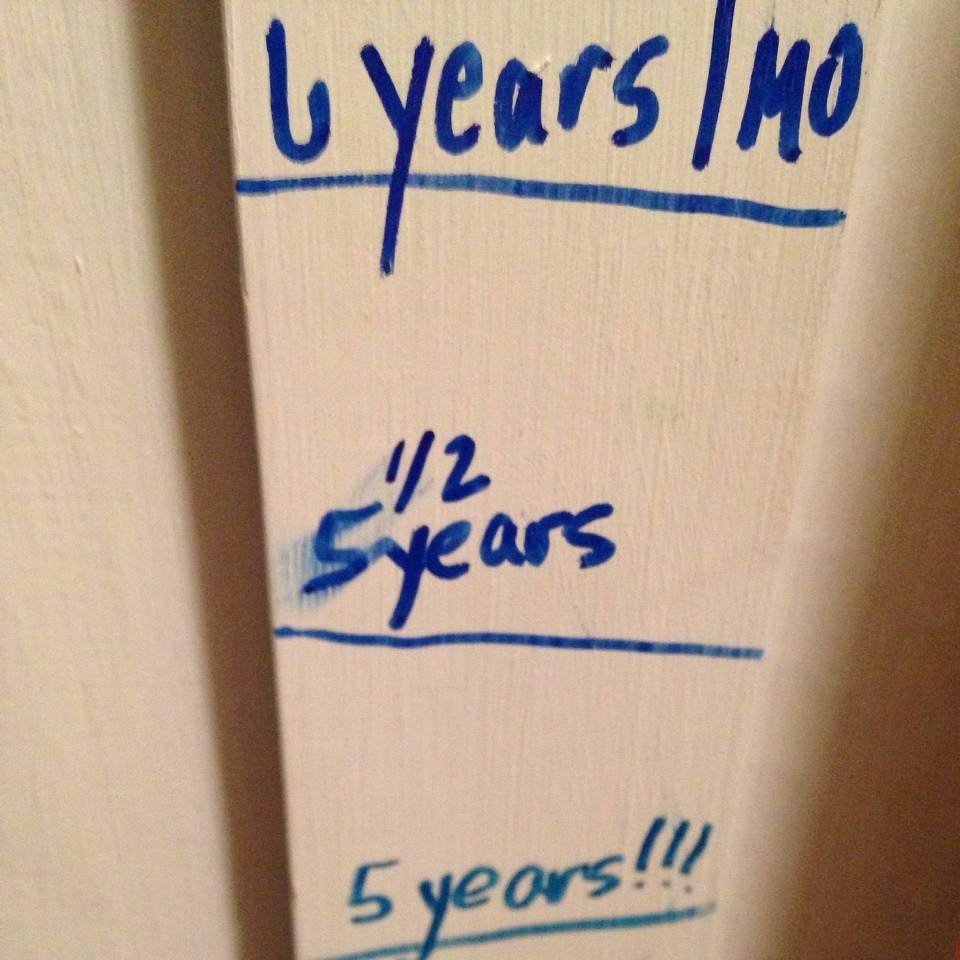 A growth chart on a wall records heights at various ages, marked with blue ink for 5 years, 5½ years, and 6 years 1 month.