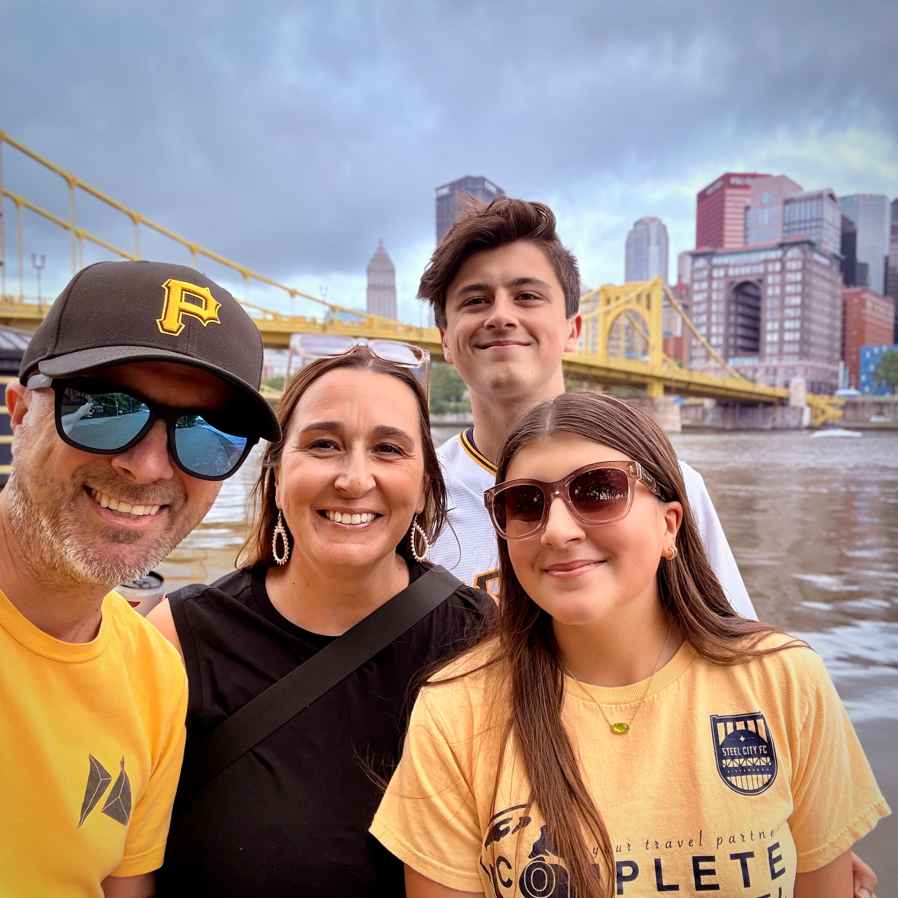 Four people are smiling together in front of a river with a cityscape and yellow bridge in the background.