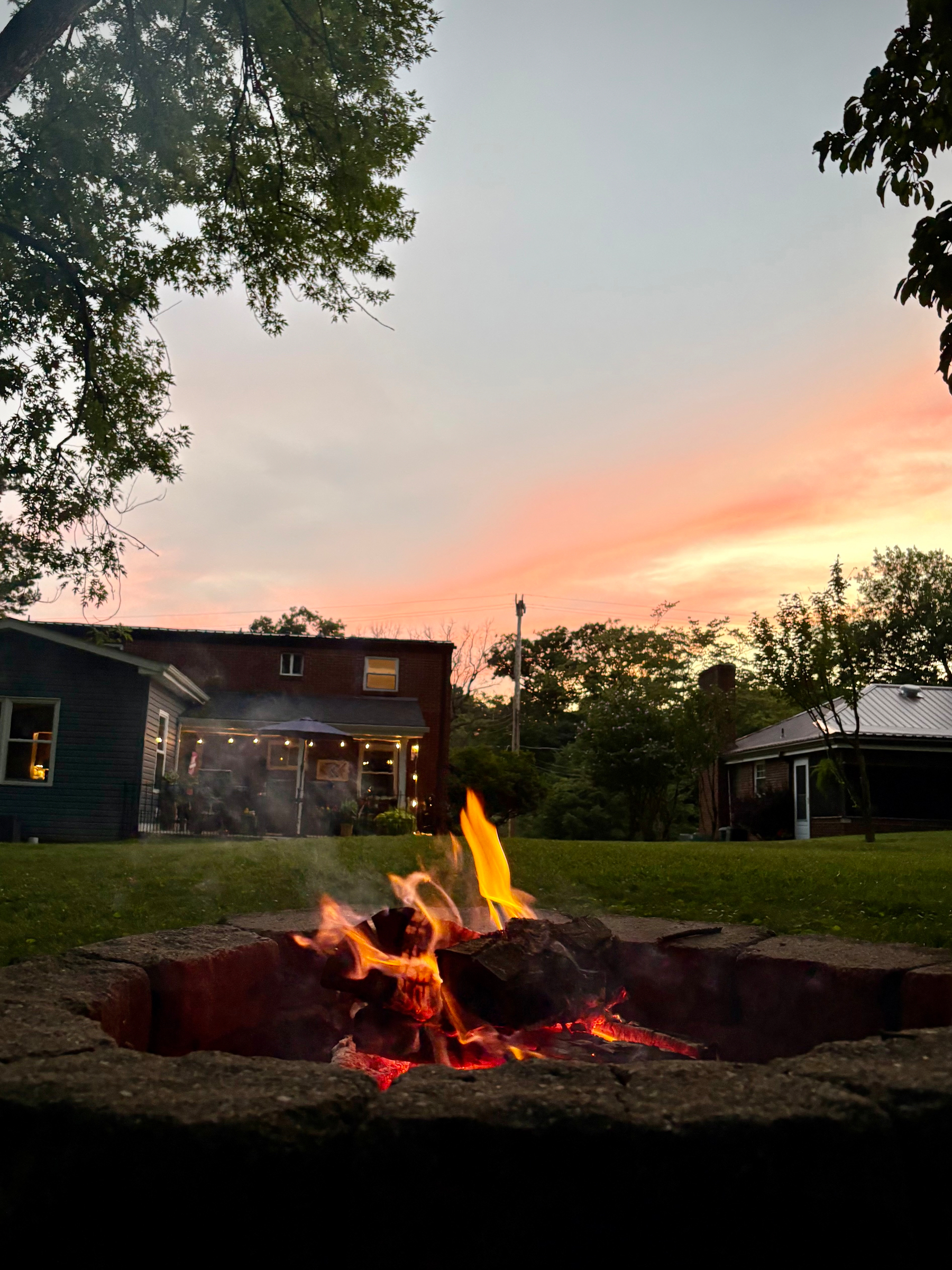 A glowing campfire burns in a stone fire pit on a grassy yard at sunset, with houses and trees in the background.