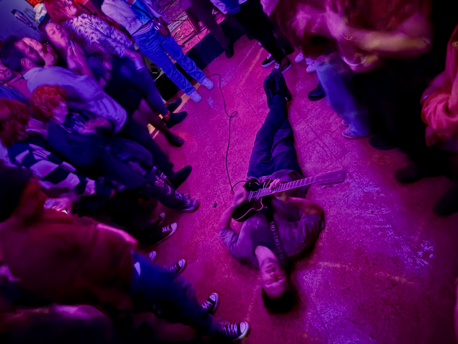 A musician is lying on the floor playing a guitar surrounded by people at a lively event.