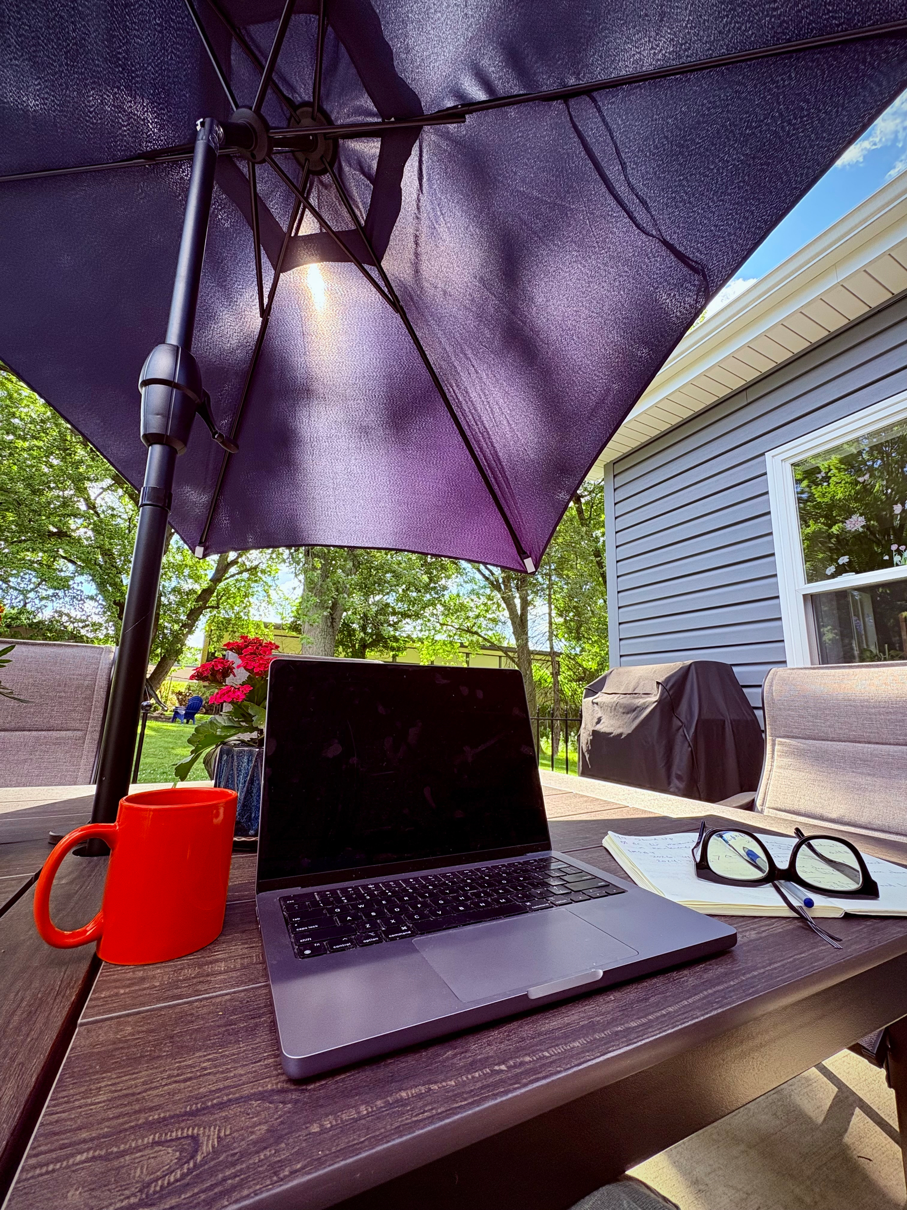 A laptop, red mug, notebook with a pen, and eyeglasses are arranged on an outdoor wooden table under a large purple patio umbrella with greenery and a house in the background.