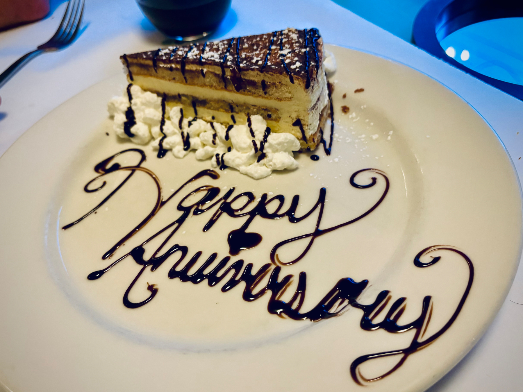 A slice of layered cake topped with chocolate drizzle and surrounded by whipped cream is served on a plate with Happy Anniversary written in chocolate syrup.