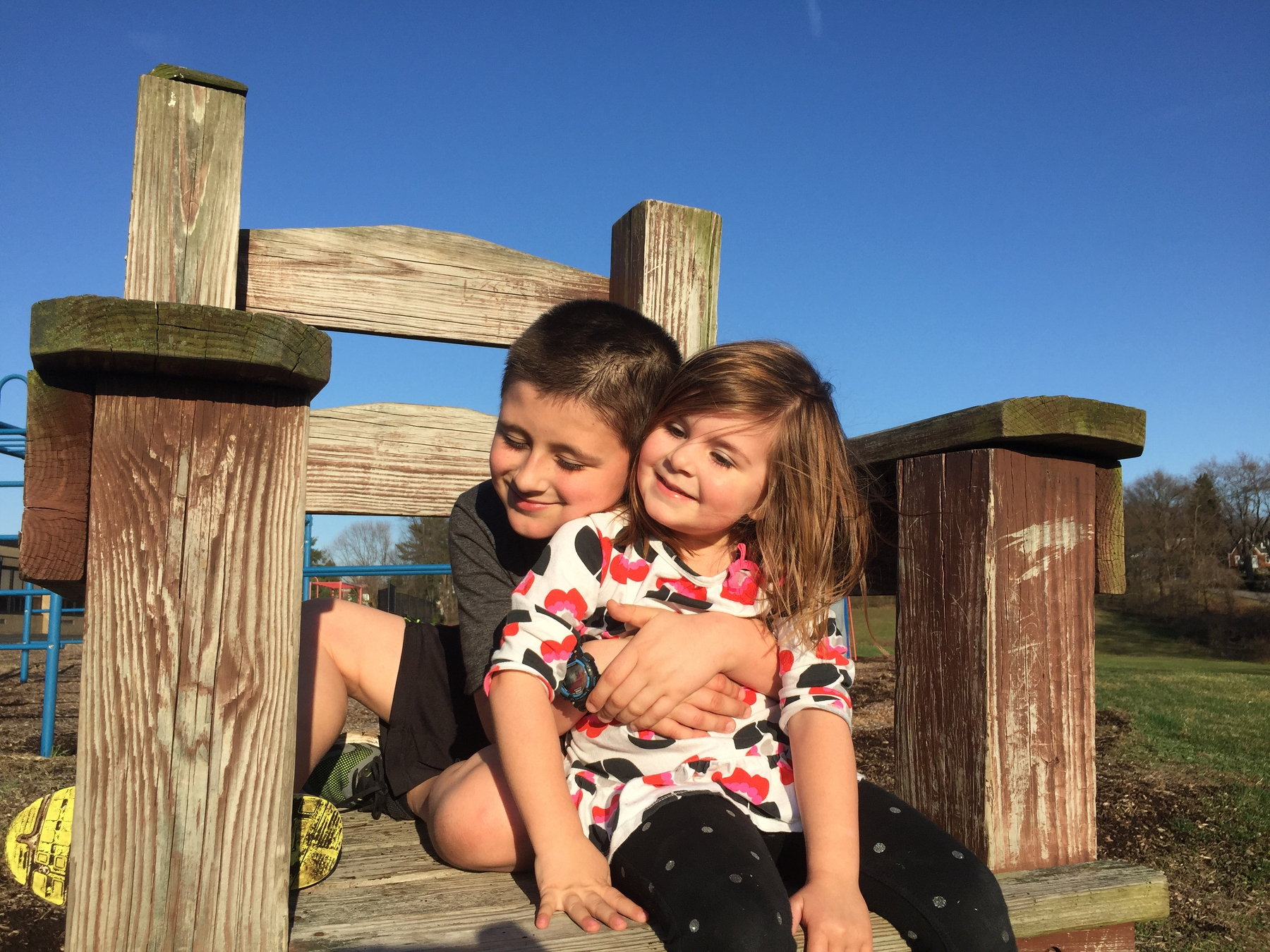 A young boy and girl are sitting on a large wooden chair, embracing each other under a clear blue sky.