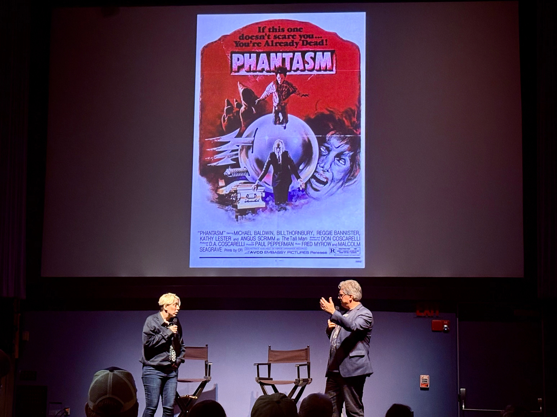 A man and a woman are standing in front of an audience on a stage with two empty chairs, discussing in front of a large screen displaying a movie poster for the film Phantasm.