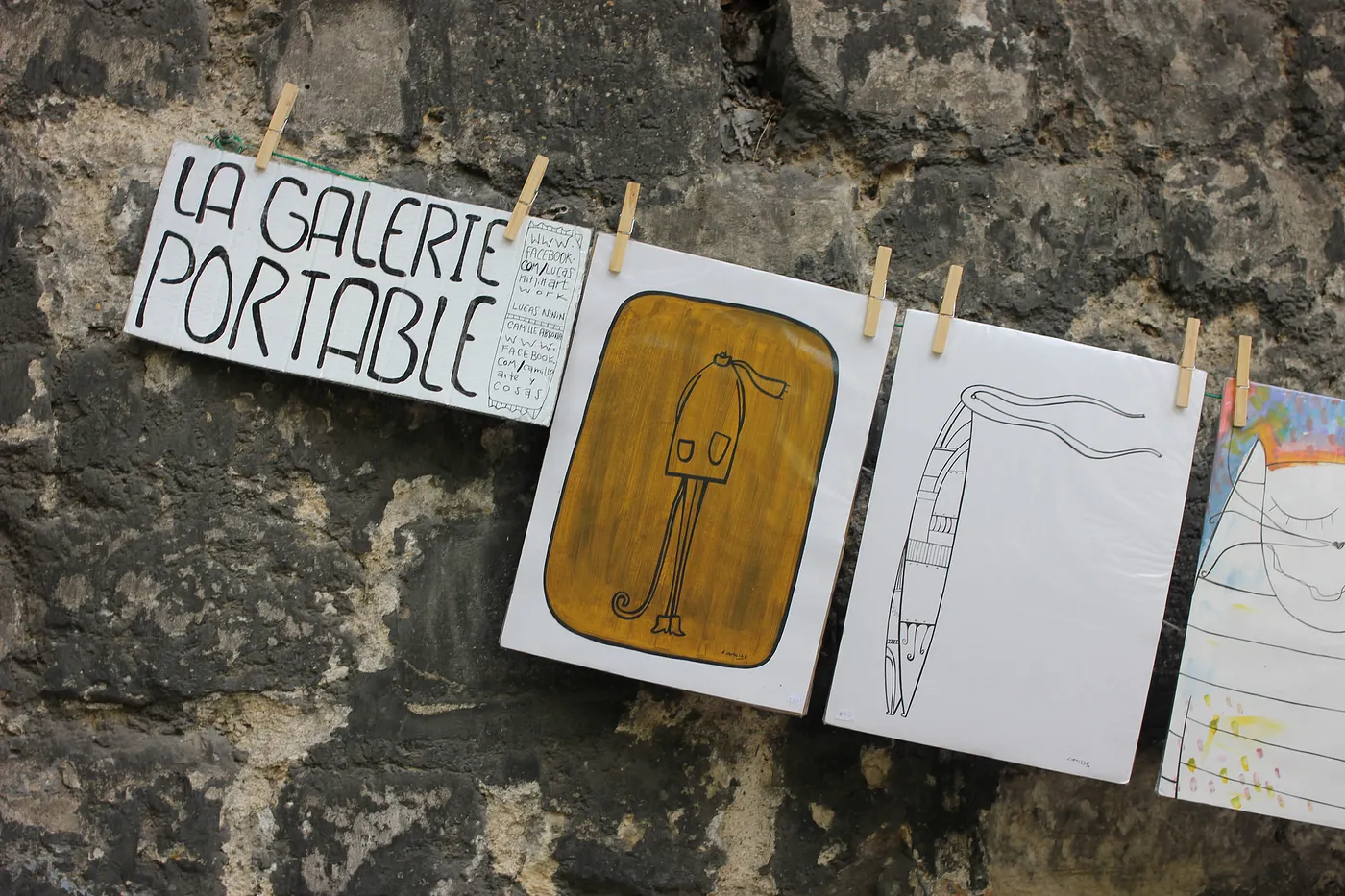 Auto-generated description: Art prints are hung on a clothesline against a stone wall, displaying various sketches and the sign La Galerie Portable.