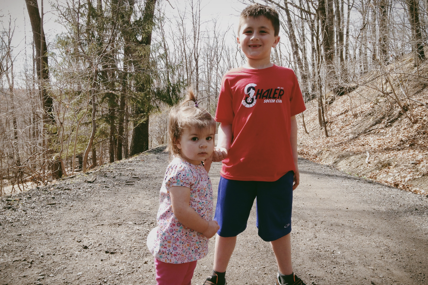 A young boy and a toddler are standing on a dirt path in a wooded area.