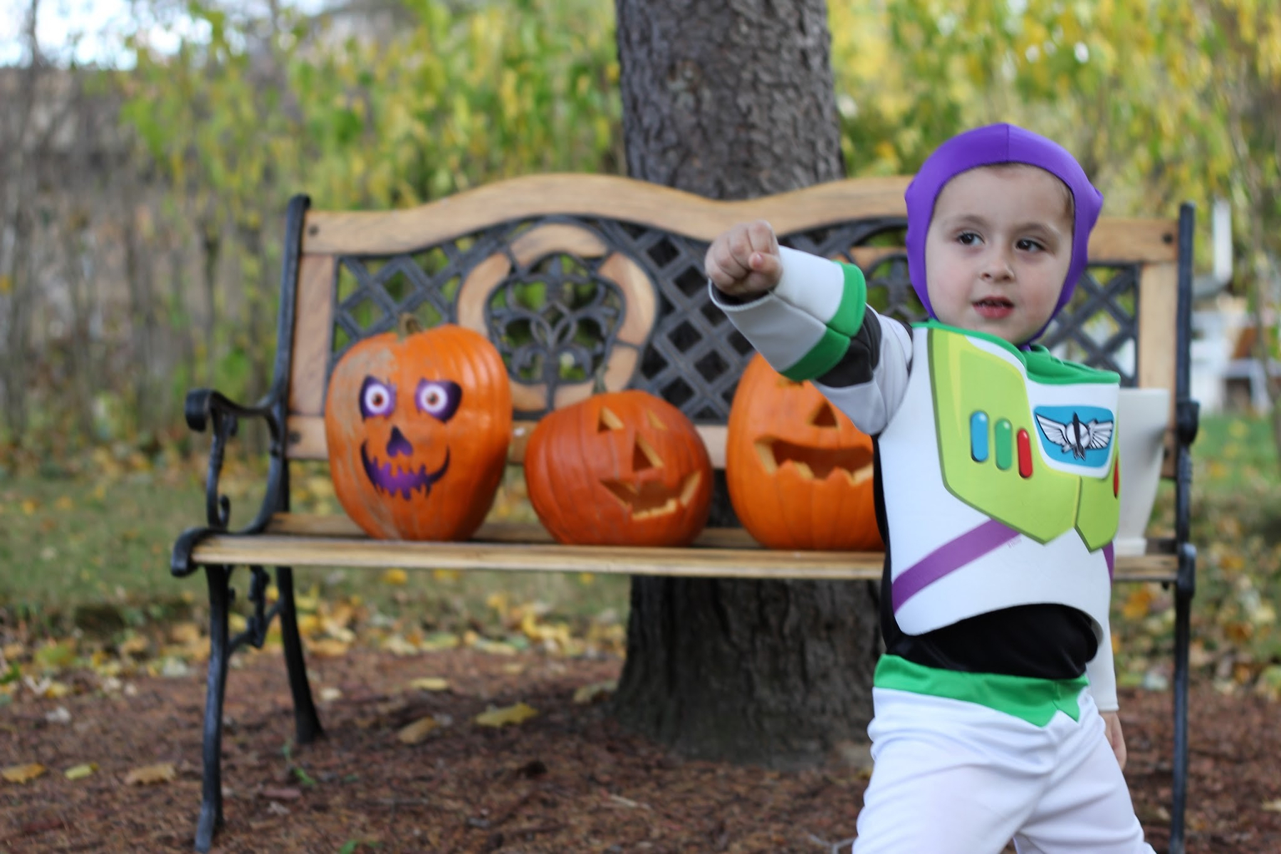 A child in a Buzz Lightyear costume poses in front of a bench with three carved pumpkins.