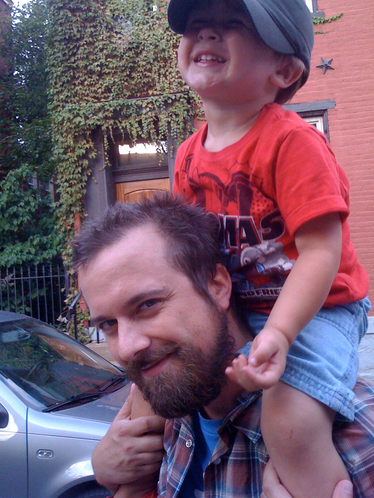 A bearded man is giving a happy child a piggyback ride on a street, with greenery and a brick building in the background.