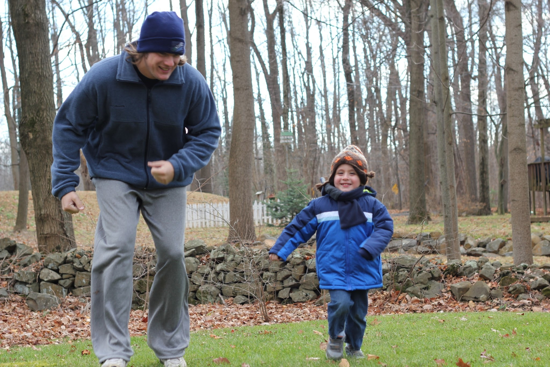A man and a child in winter clothing are joyfully running outdoors on grass surrounded by trees.