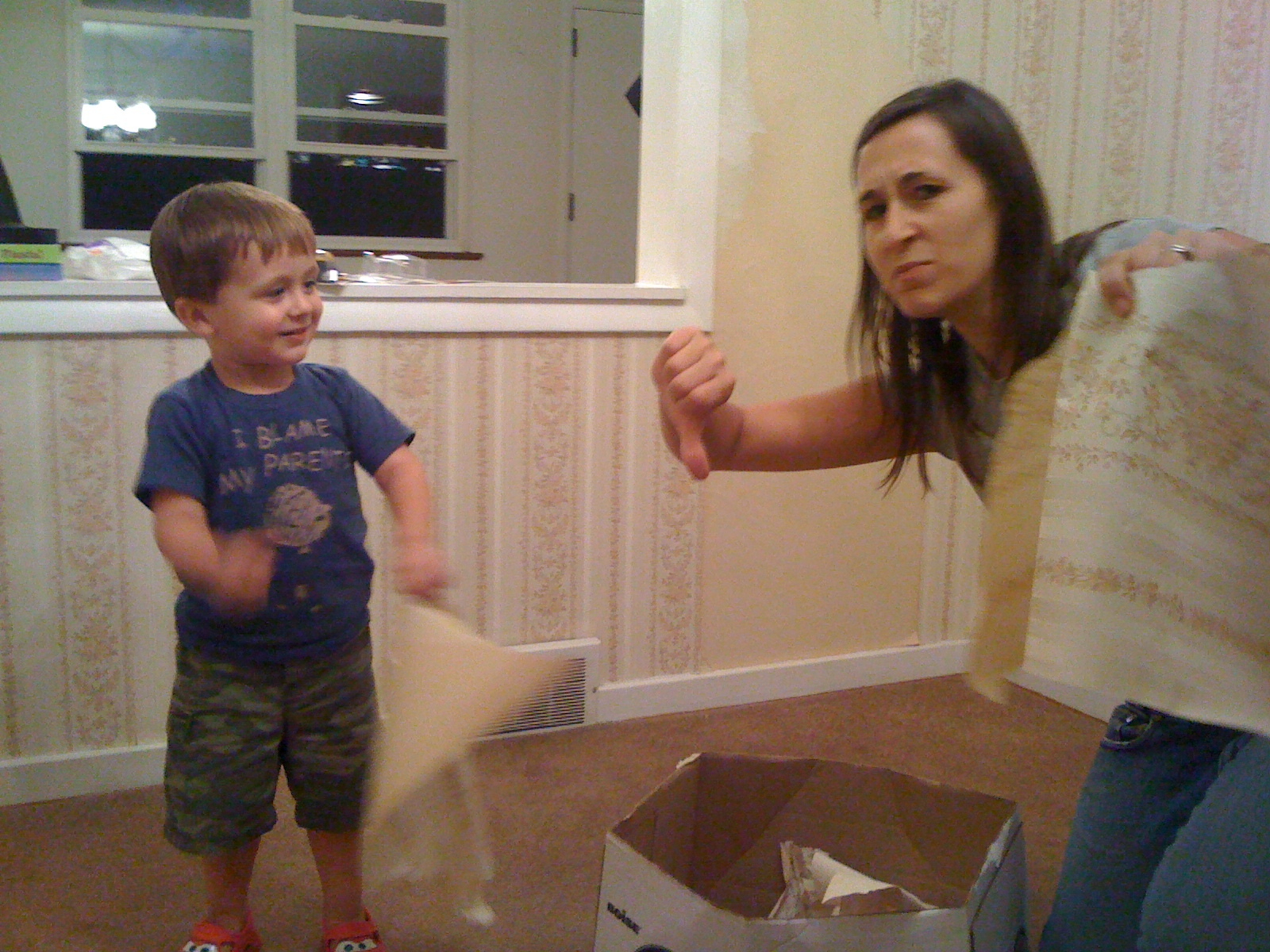 A young boy and a woman are in a room with wallpaper and appear to be engaged in a playful disagreement, with the woman giving a thumbs-down gesture while holding wallpaper pieces.