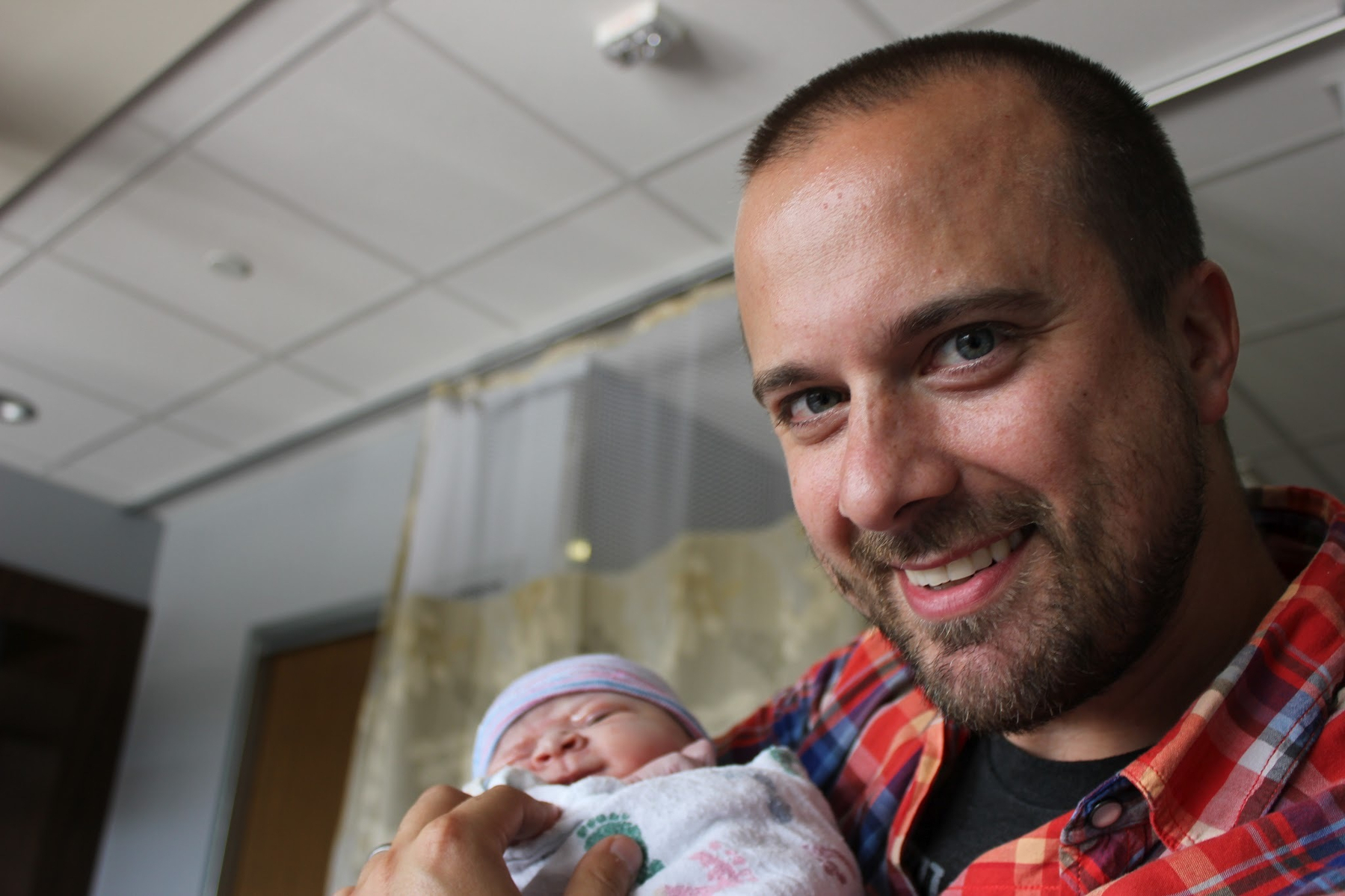 A smiling man holds a swaddled newborn in his arms, indoors.