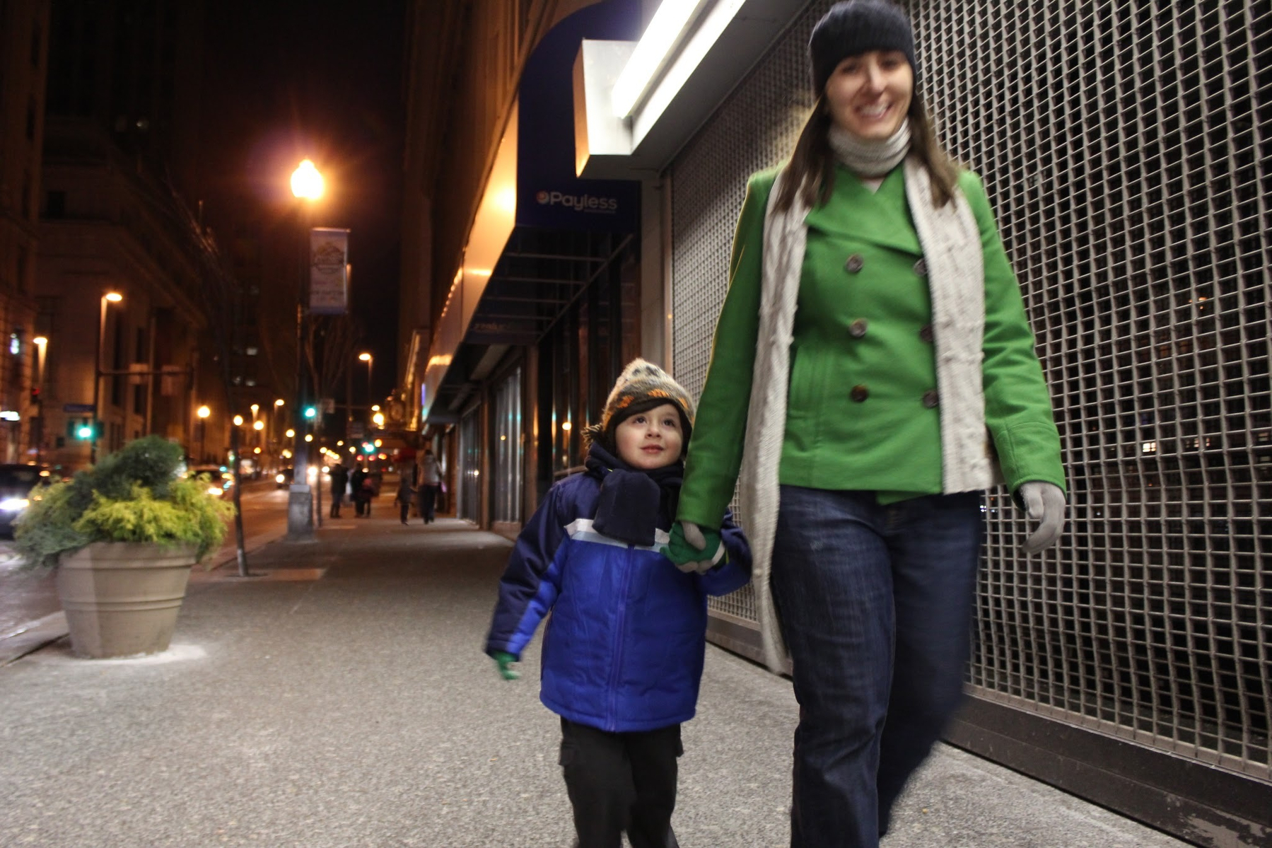 A woman and a child are walking hand-in-hand on a city sidewalk at night, dressed in winter clothing.