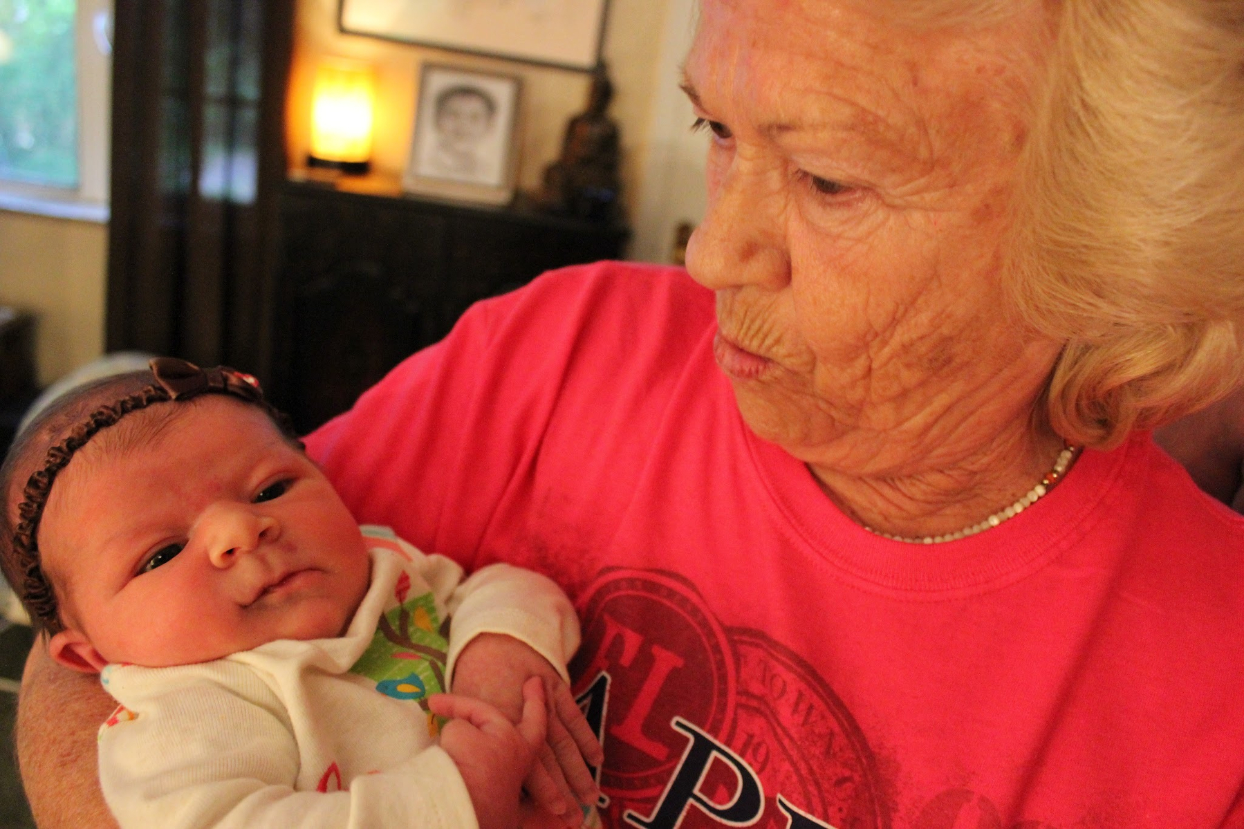 An older woman is holding a baby dressed in a white outfit with a decorative headband.