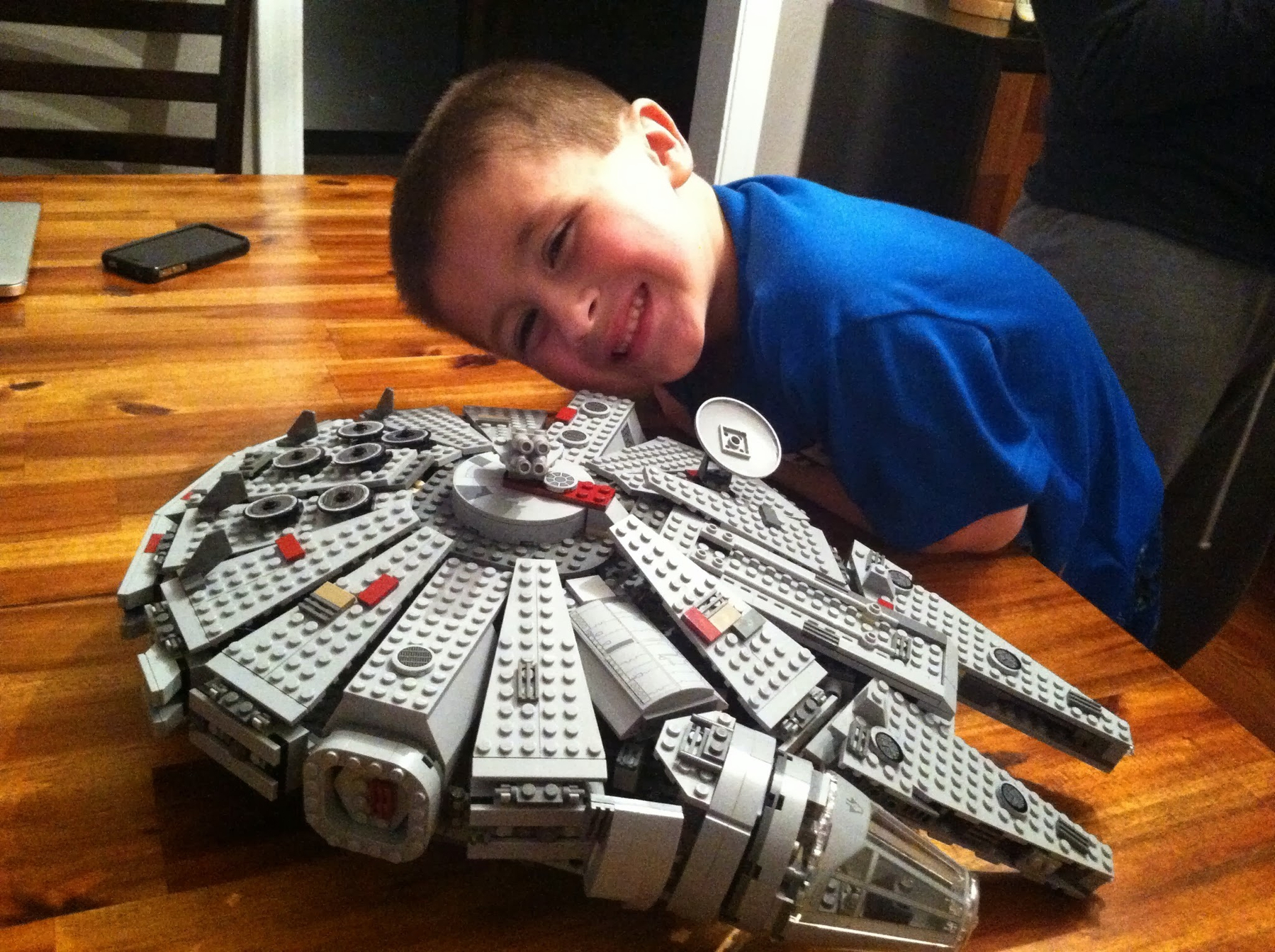 A young boy happily poses with a detailed LEGO model of the Millennium Falcon on a wooden table.