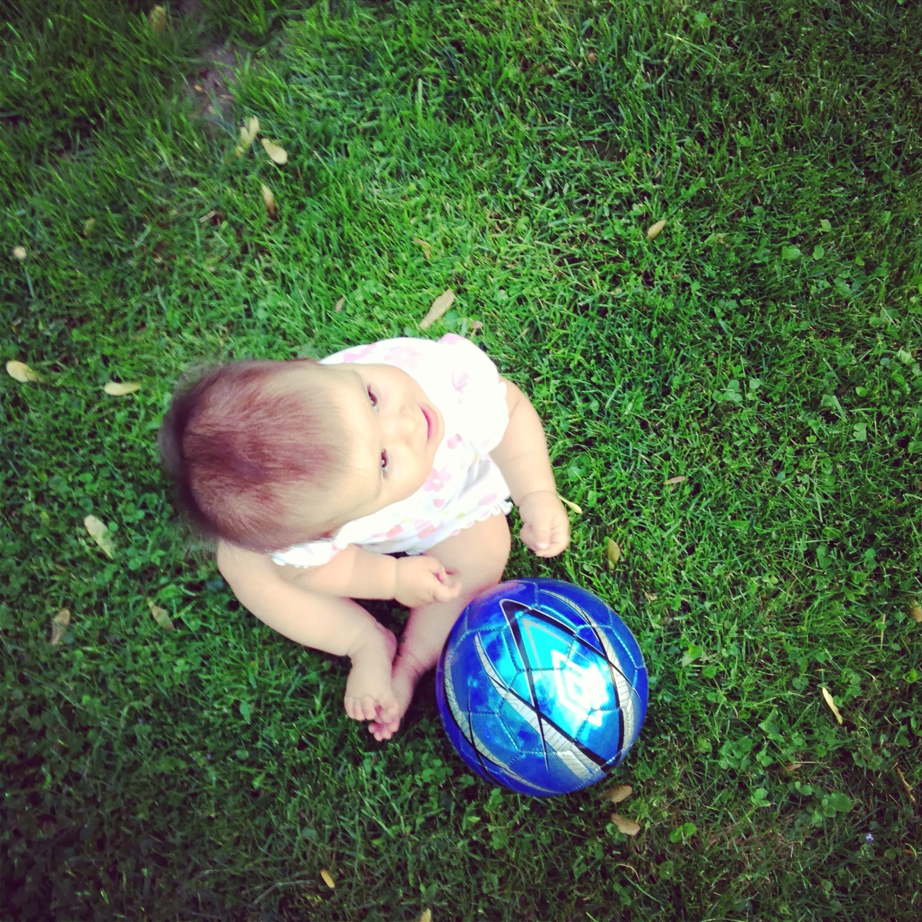 A baby is sitting on grass next to a blue soccer ball.