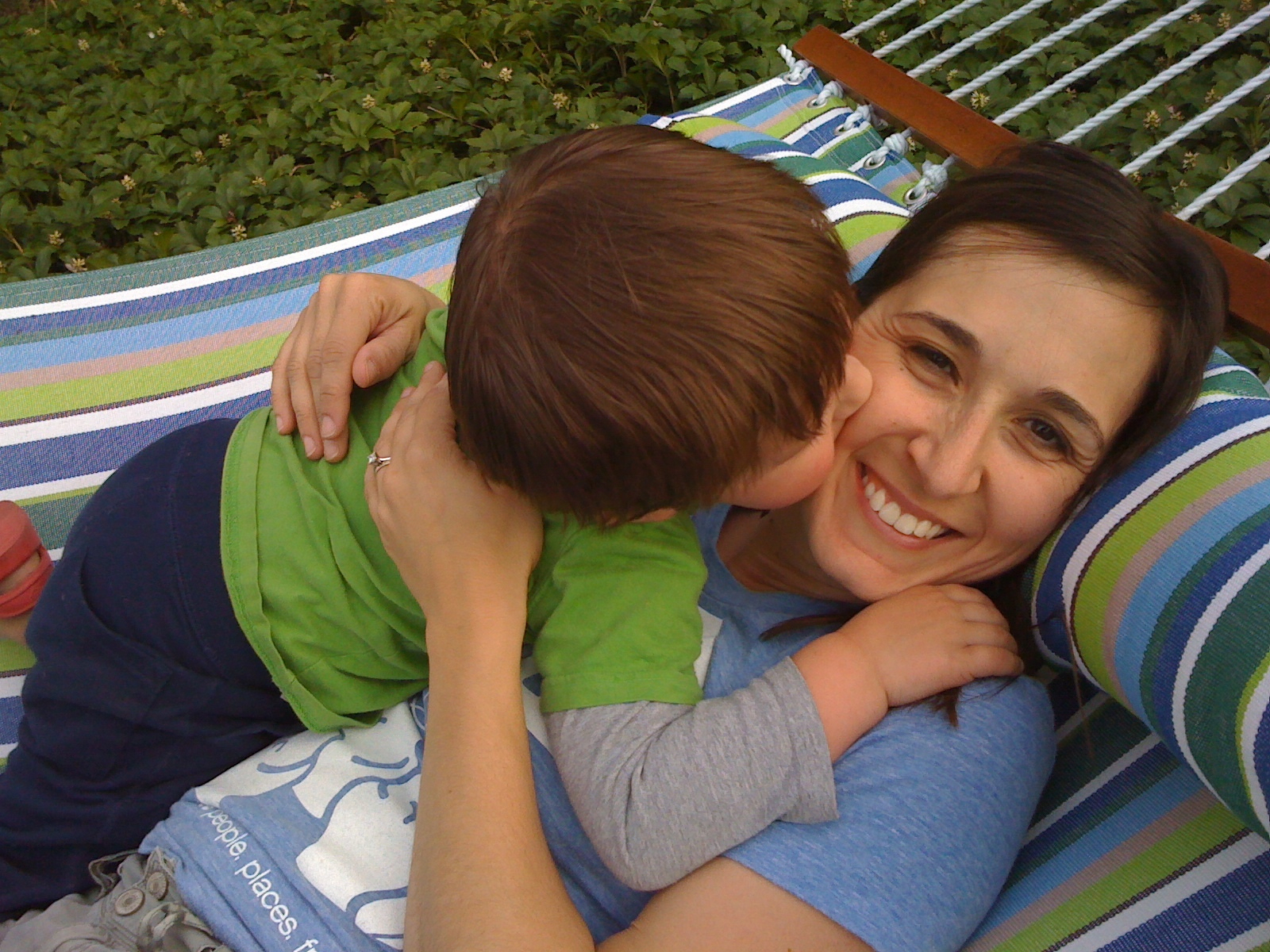 A woman is smiling while a child leans over and gives her a kiss on the cheek as they relax on a striped hammock.