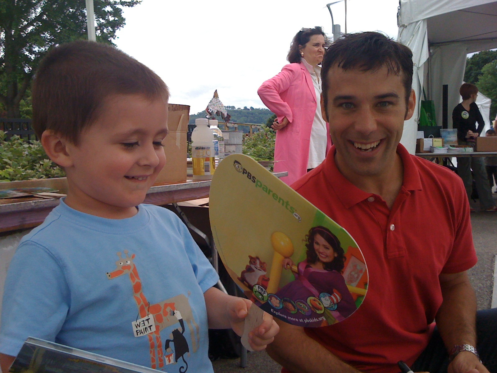 A smiling man and a young boy are sitting outdoors under a tent, interacting happily while the boy holds a colorful brochure.