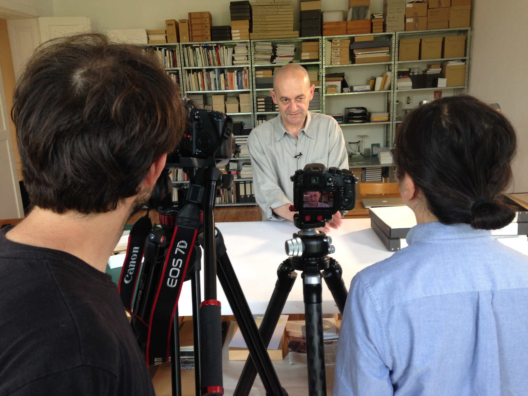 Auto-generated description: Two people are setting up cameras to photograph or film a man standing by a table in a room full of shelves with various items.