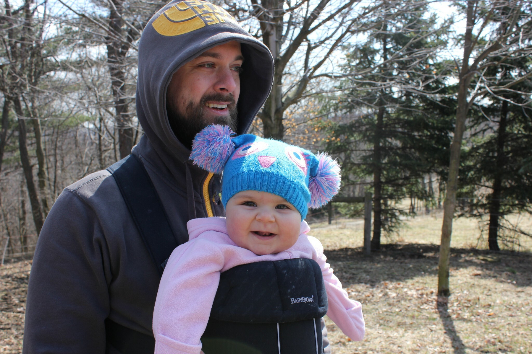 A person is carrying a smiling baby in a blue hat with bear ears while standing in a park.