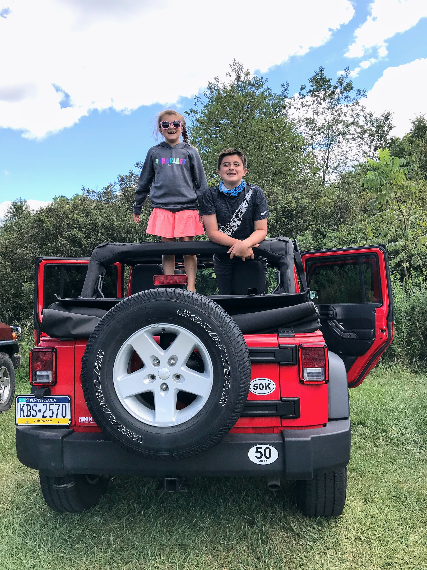 Two children are happily standing and sitting on the back of a red Jeep parked on grass with trees and a partly cloudy sky in the background.