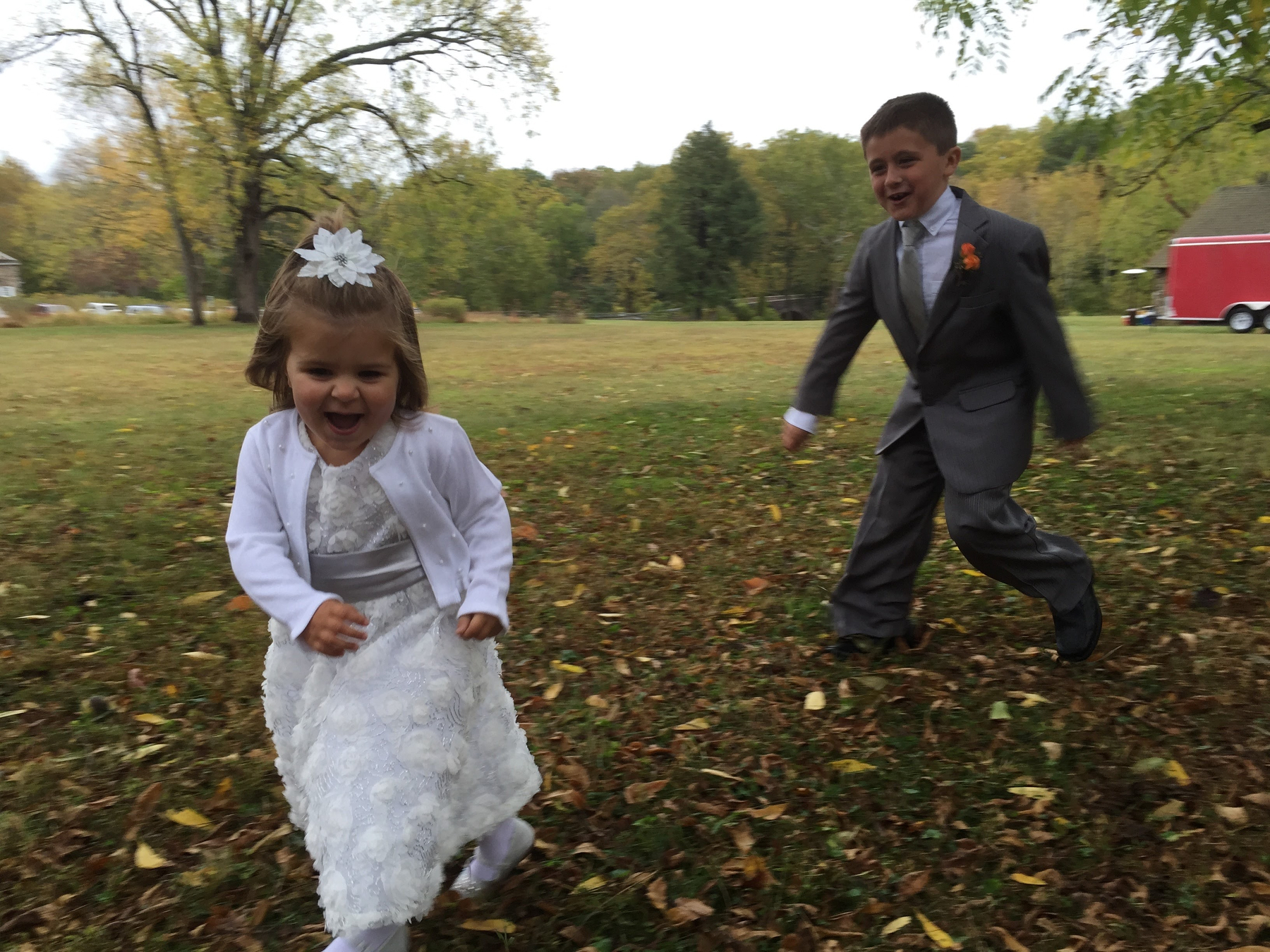 Two children in formal attire are joyfully running on a grassy area surrounded by trees.
