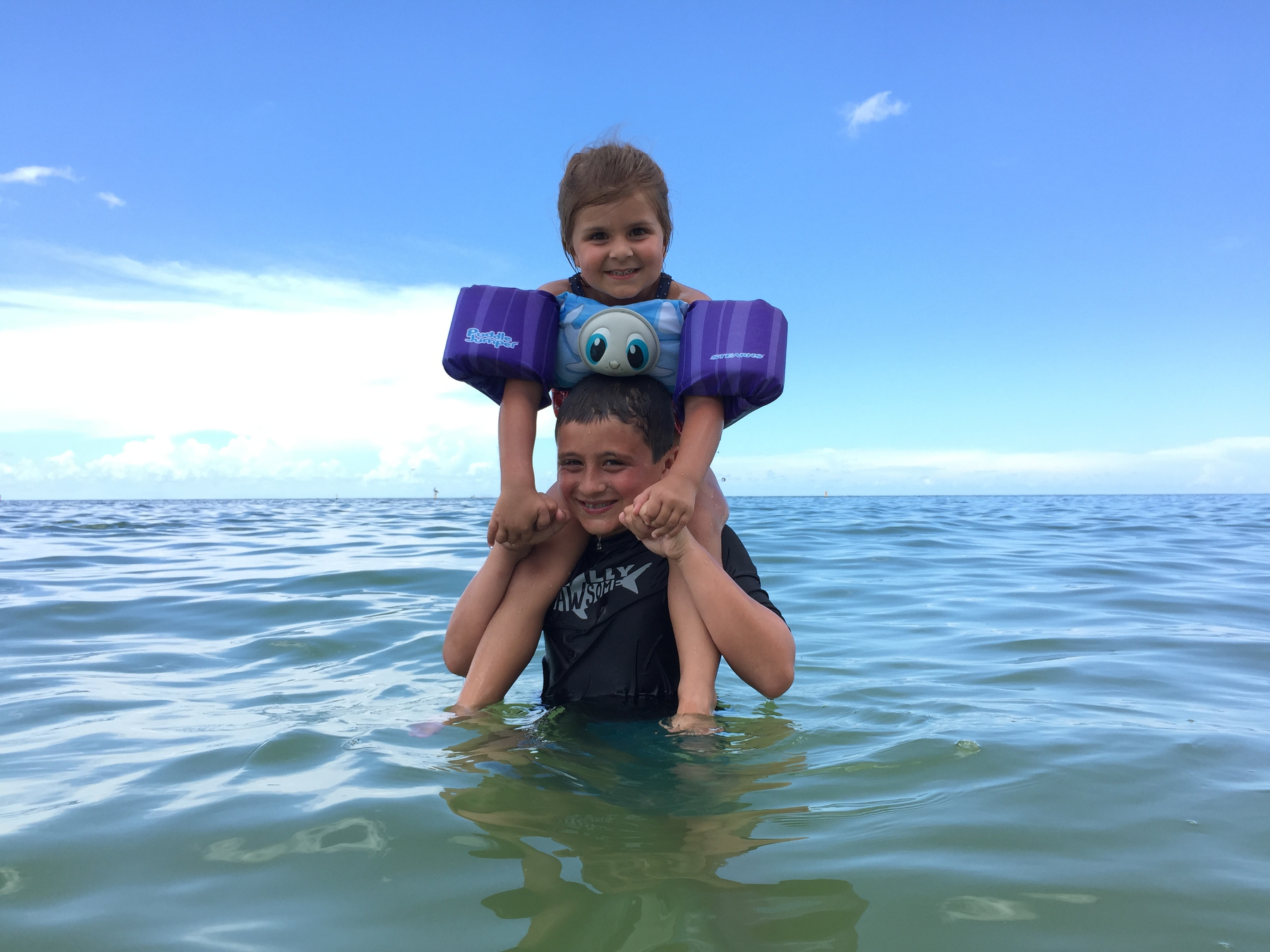 A child wearing floaties is sitting on someone's shoulders in the ocean, under a clear blue sky.
