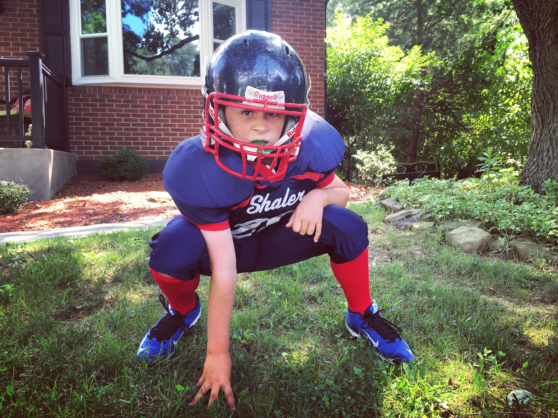 A child wearing a football helmet and uniform is posing in a crouched stance on a grassy area in front of a house.