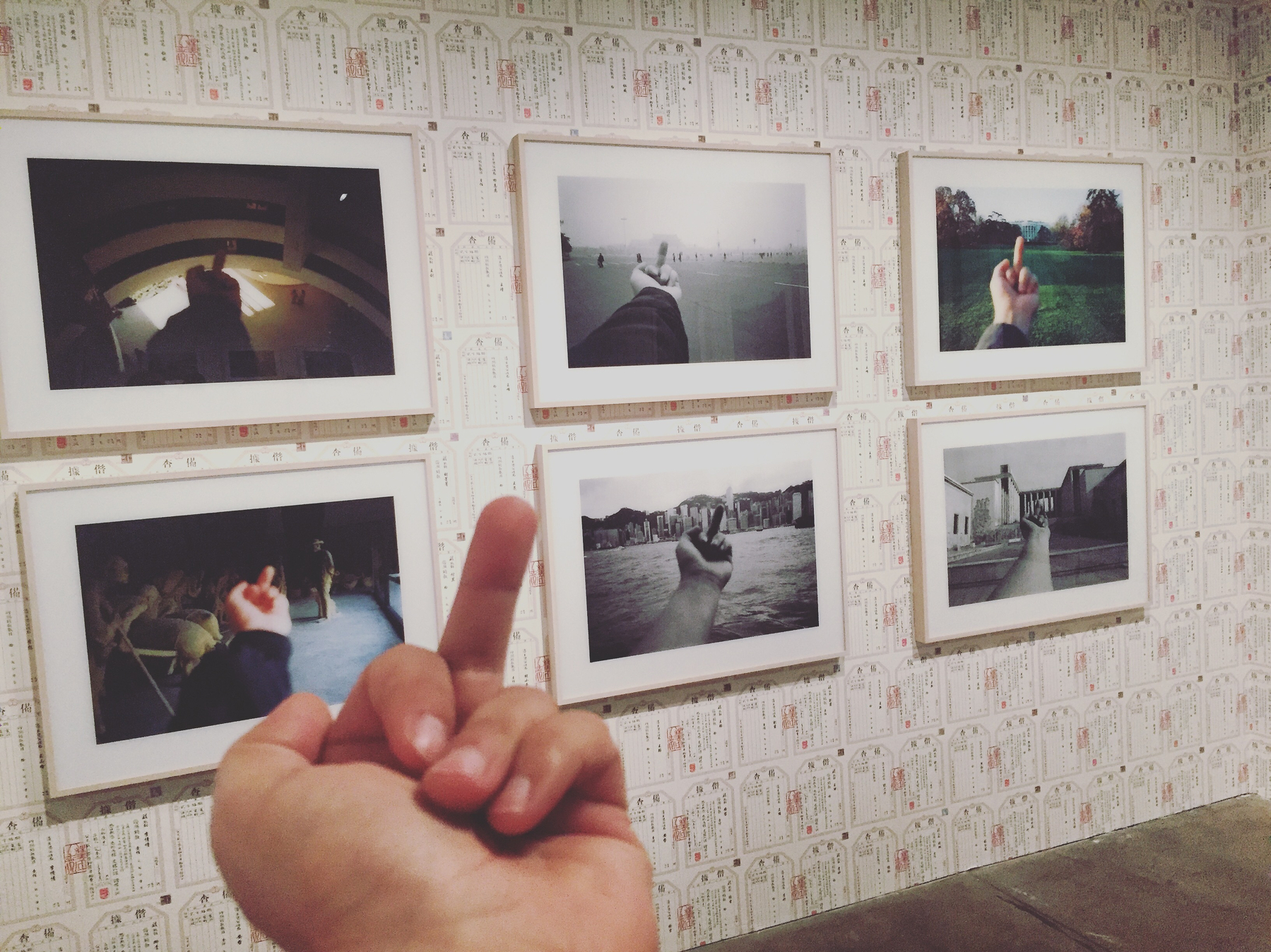 A hand making a rude gesture is seen in front of a gallery wall displaying framed photographs of similar gestures.