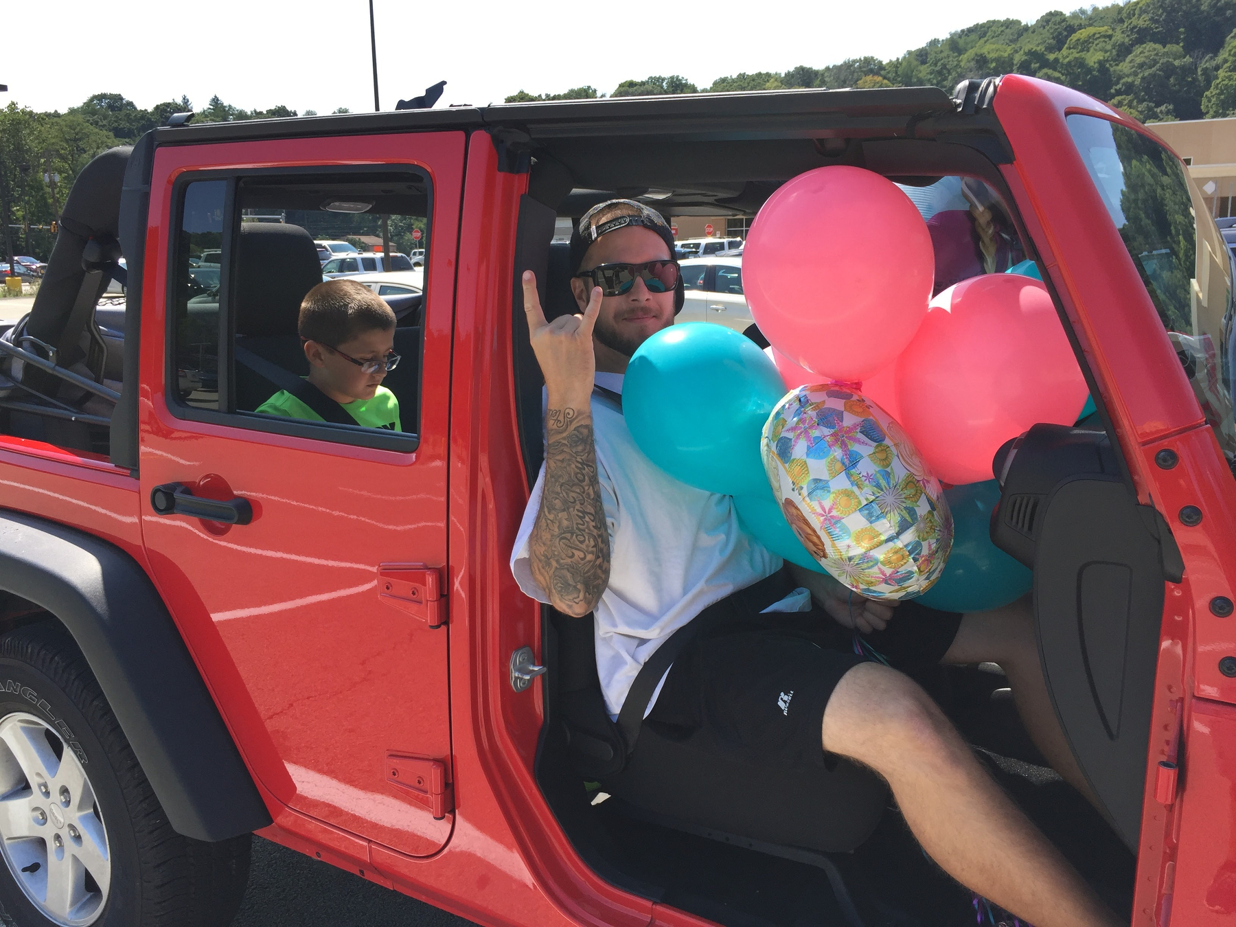 A man holding colorful balloons and making a hand gesture is sitting in the driver's seat of a red jeep, while a boy is seated in the back.