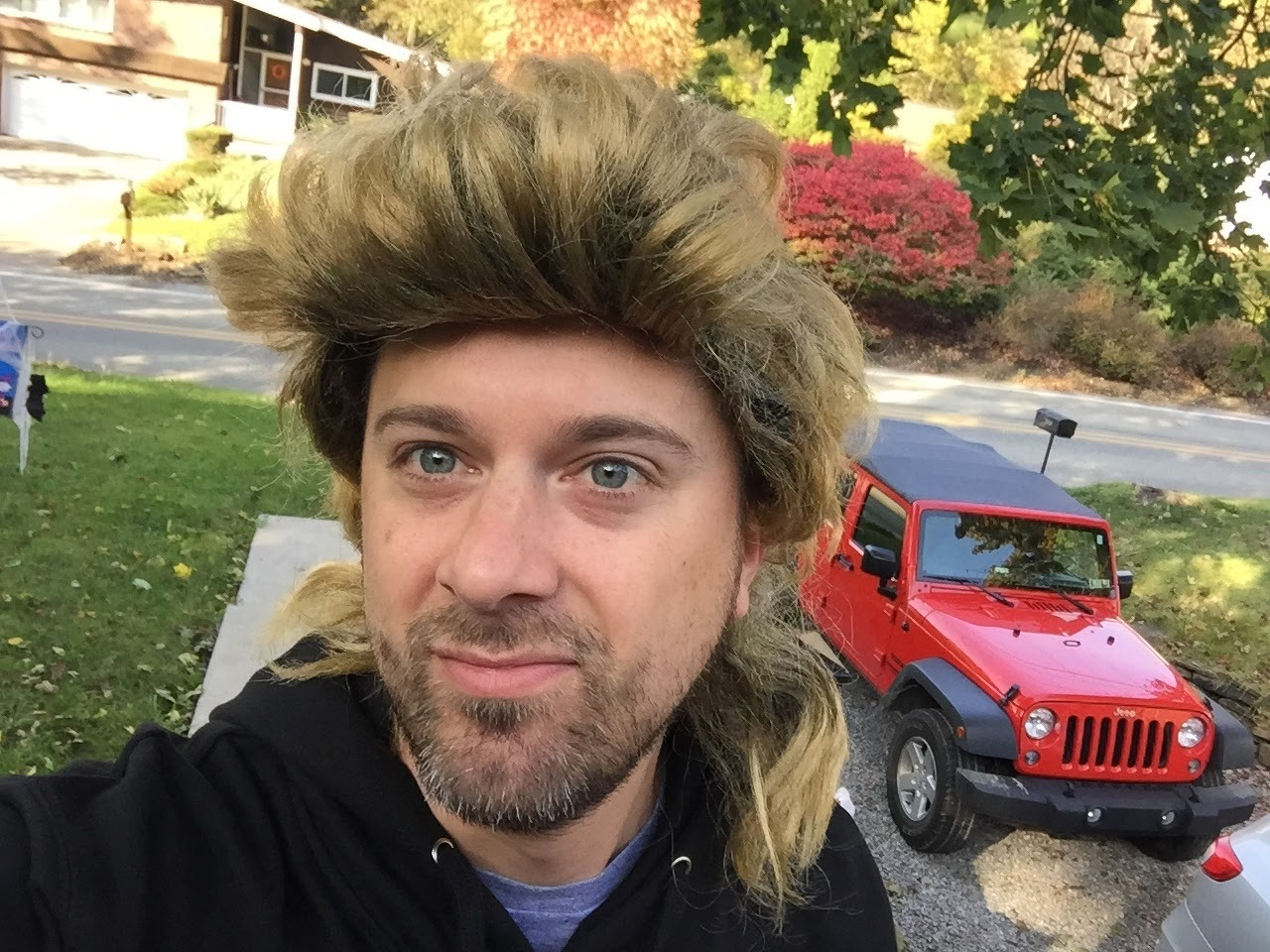 A man wearing a blonde mullet wig and a black hoodie is taking a selfie outdoors with a red Jeep parked in the background.