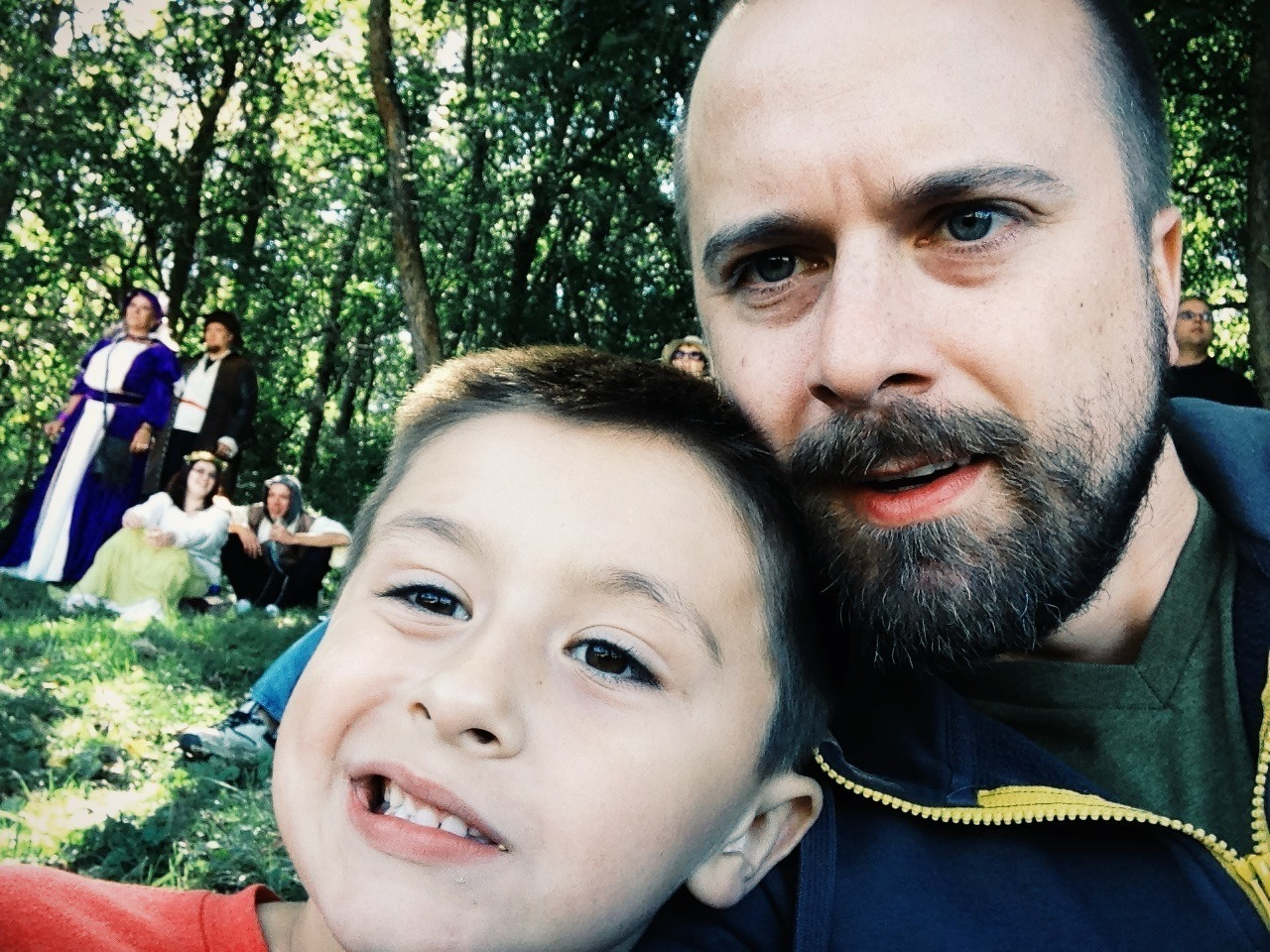 A man and a child take a selfie outdoors with people dressed in historical costumes in the background.